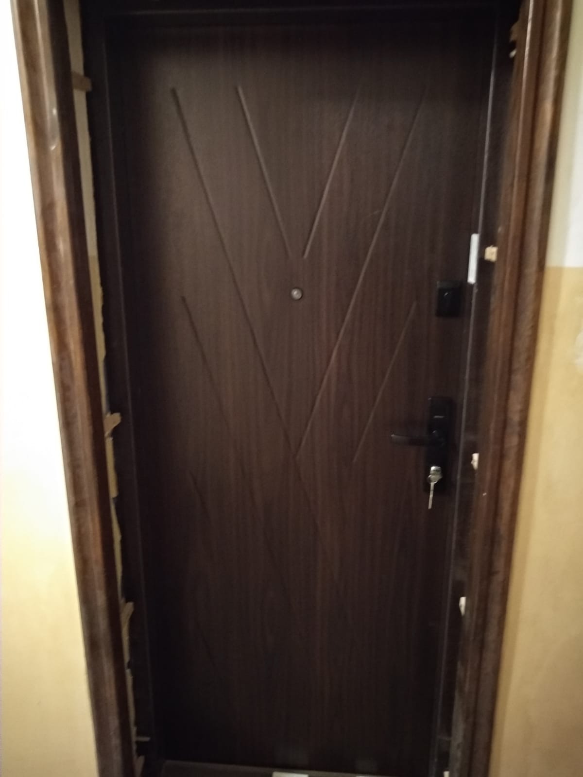 Ciemnobrązowe drzwi wejściowe z geometrycznym wzorem w kształcie litery V, widoczny zamek z kluczem i uszkodzona ościeżnica.