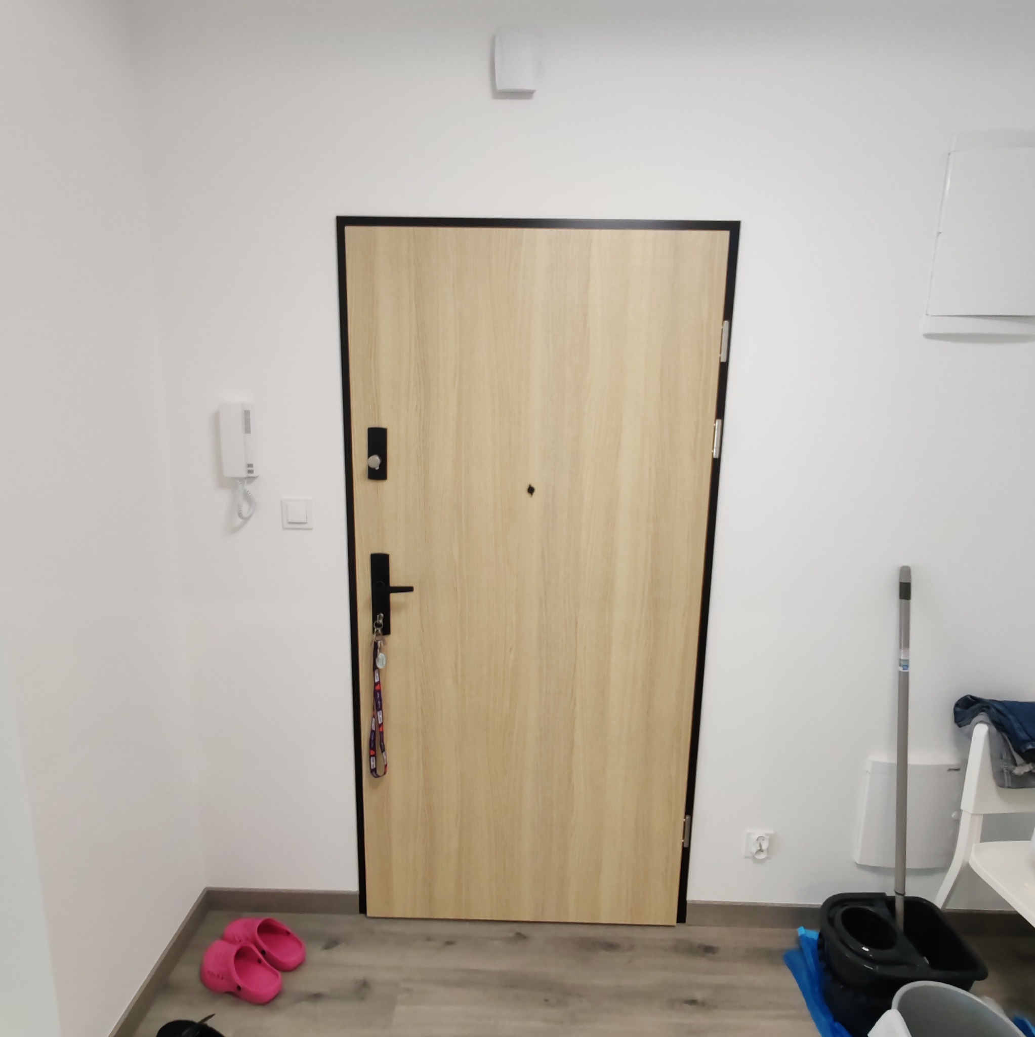 Jasne, drewnopodobne drzwi wejściowe z czarną ramą i okuciami, widoczne od wewnątrz mieszkania, z kluczami na klamce, w tle białe ściany i mop z wiadrem.