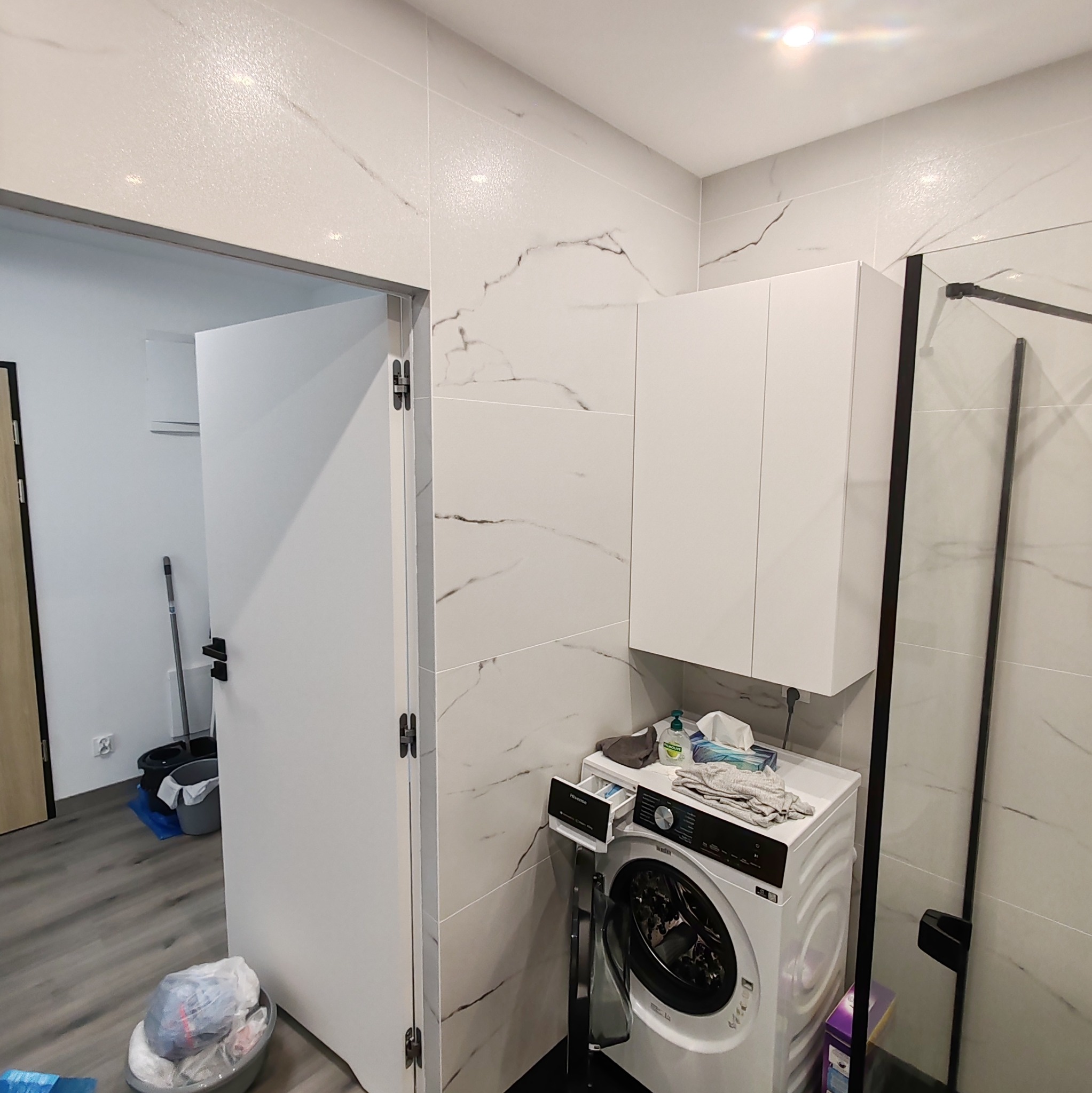 Nowoczesna łazienka z białą pralką, szafką wiszącą i kabiną prysznicową z czarnymi profilami, wykończona płytkami imitującymi marmur.