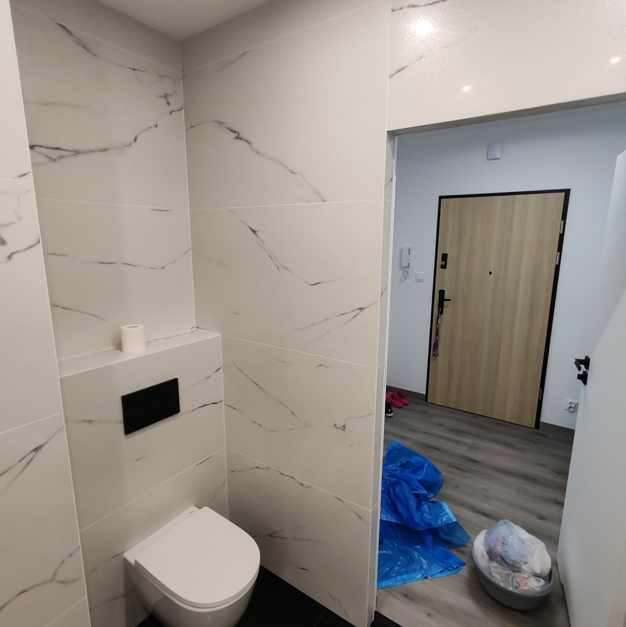 Nowoczesna toaleta z białymi płytkami imitującymi marmur, wisząca miska WC z czarnym przyciskiem, widok na przedpokój z brązowymi drzwiami i workiem na śmieci.