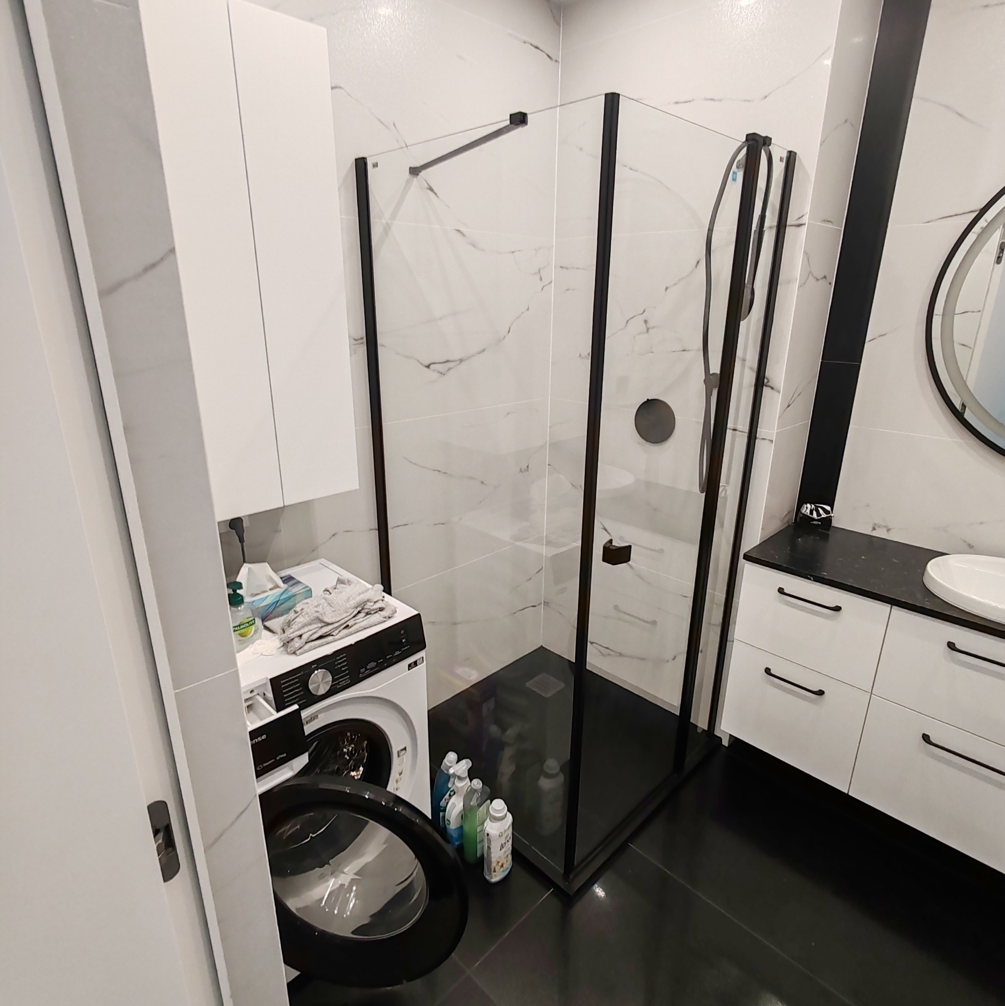 Nowoczesna łazienka z kabiną prysznicową typu walk-in z czarnymi profilami, białą umywalką z szafką i pralką z otwartymi drzwiami, wykończona białymi płytkami imitującymi marmur i czarną podłogą.