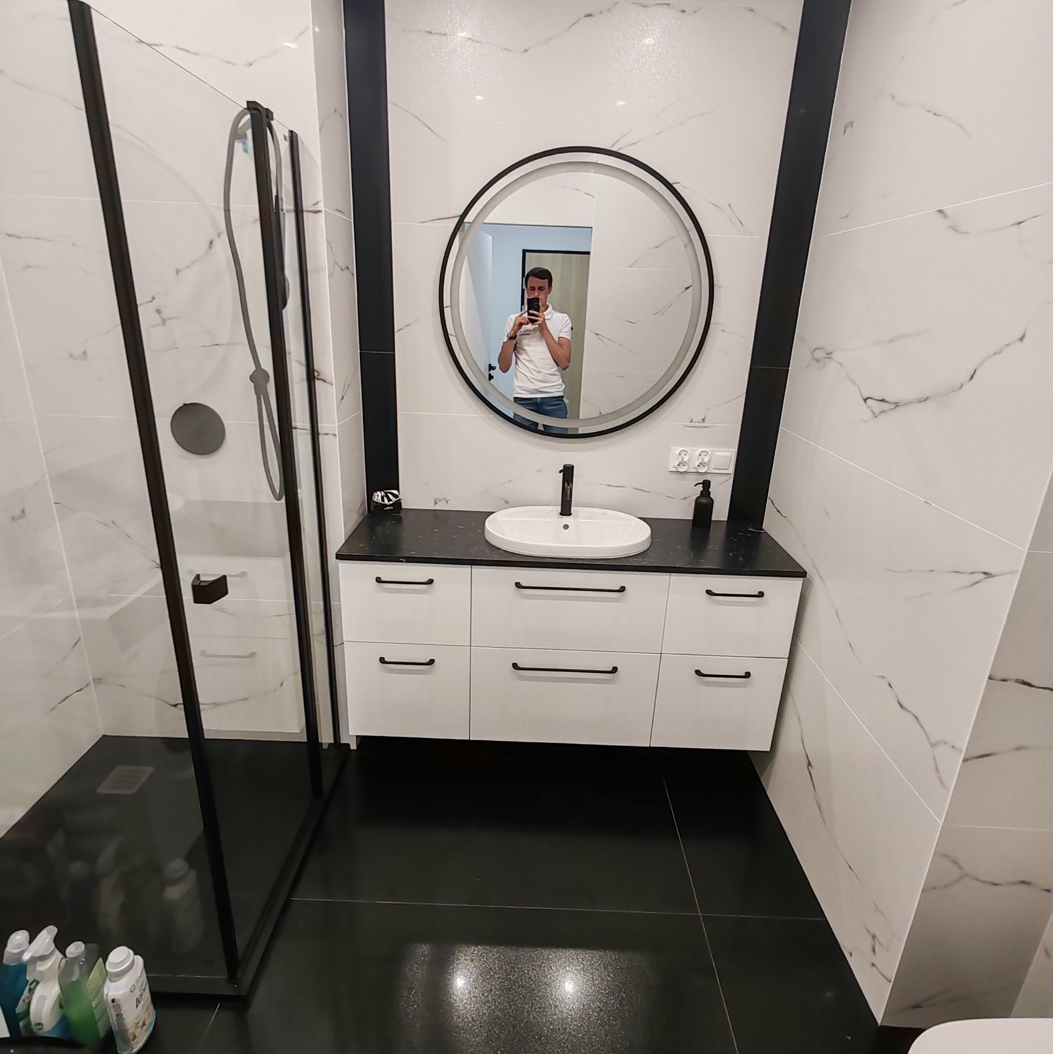 Nowoczesna łazienka z kabiną prysznicową typu walk-in, okrągłym lustrem z podświetleniem, białą szafką pod umywalką z czarnym blatem i czarnymi akcentami, wykończona białymi płytkami imitującymi...