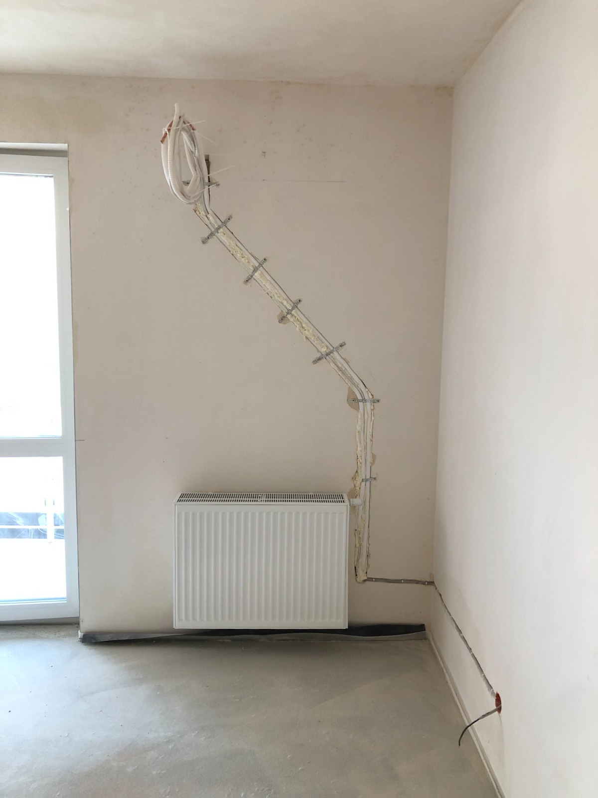 Surowy stan wnętrza: grzejnik, okno PCV i instalacja grzewcza przymocowana do ściany, zabezpieczona pianką montażową, widoczne kable elektryczne.