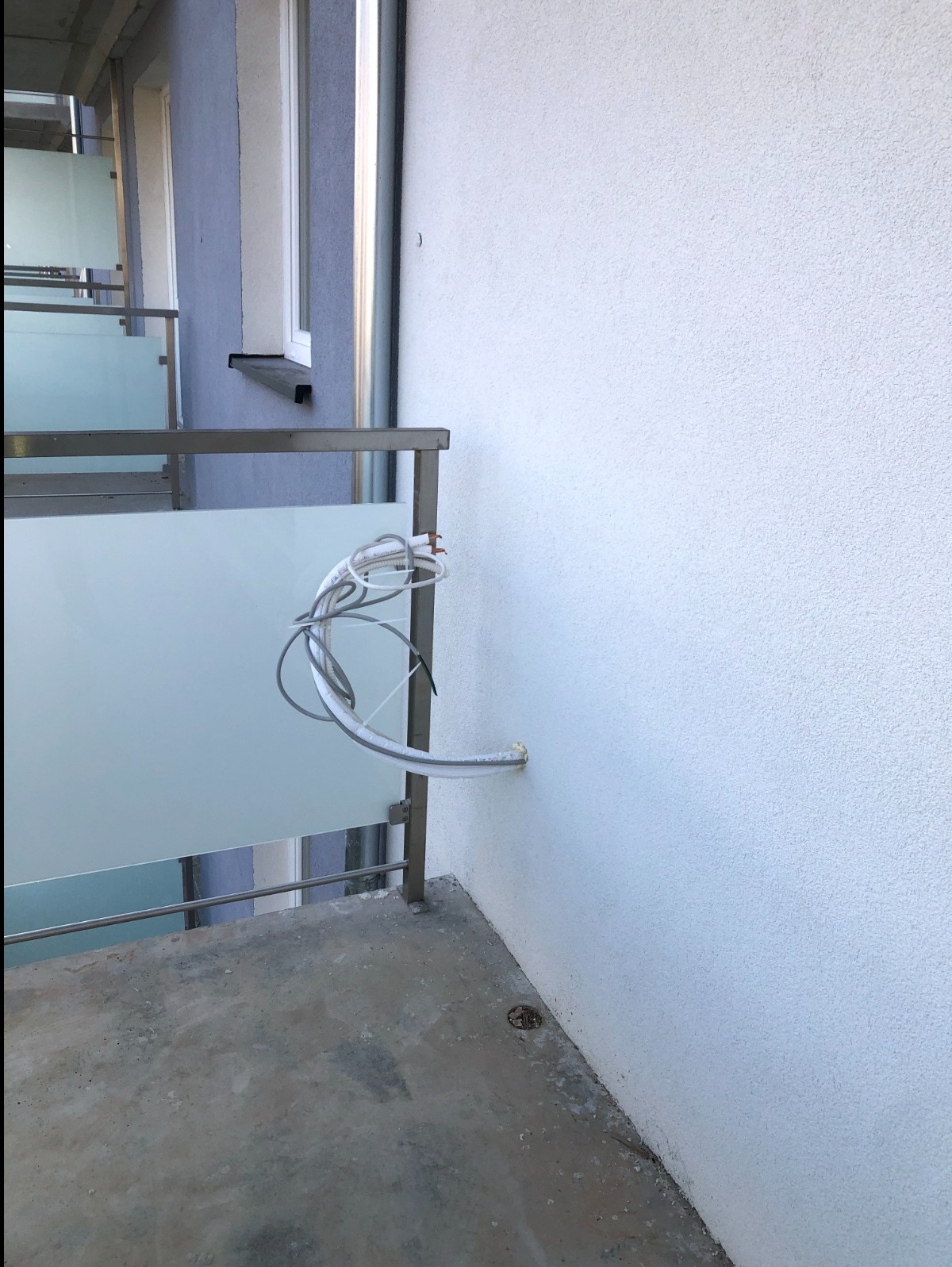 Biała ściana budynku z wystającymi kablami klimatyzacji, widoczne elementy balkonu z matowym szkłem i metalową konstrukcją.