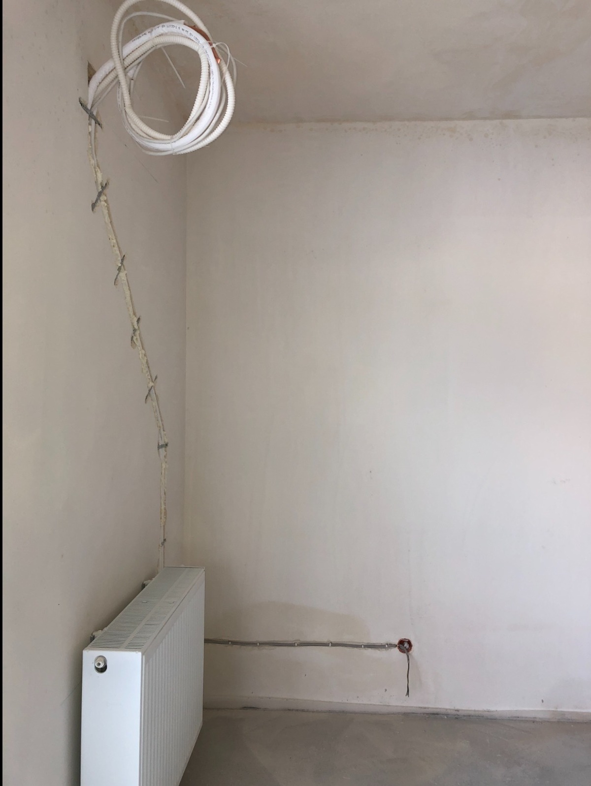 Surowy stan wnętrza: biały grzejnik, wystające kable elektryczne w białej osłonie przymocowane do ściany, gładzie na ścianach w trakcie prac wykończeniowych, widoczne ślady po instalacji elektrycznej.