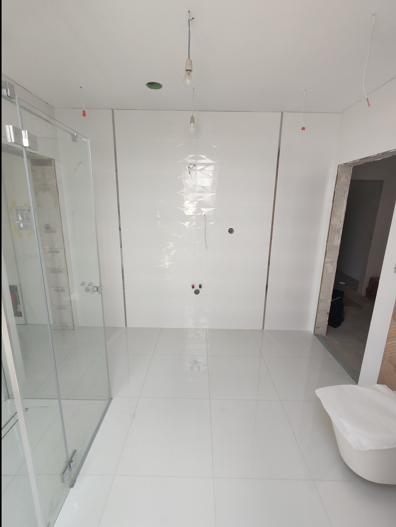 Wnętrze łazienki w trakcie remontu, widoczne białe płytki na ścianach z dekoracyjnym wzorem, szklana kabina prysznicowa, podłączenia hydrauliczne oraz wiszące kable oświetleniowe.