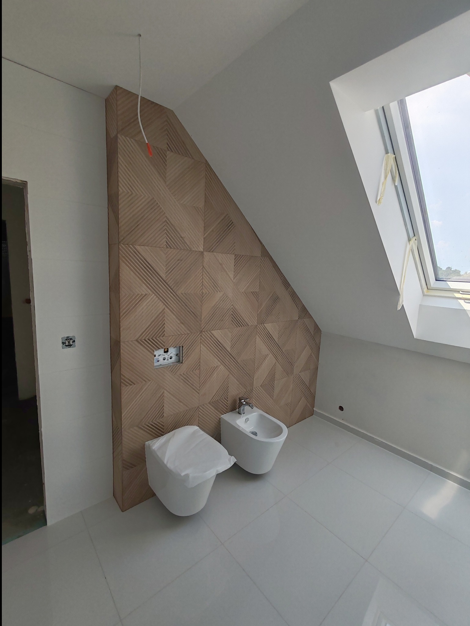 Nowoczesna łazienka na poddaszu ze skośnym oknem dachowym, drewnianą ścianą z geometrycznym wzorem, białą ceramiką sanitarną (toaleta zakryta folią ochronną i bidet) oraz jasnymi, błyszczącymi...