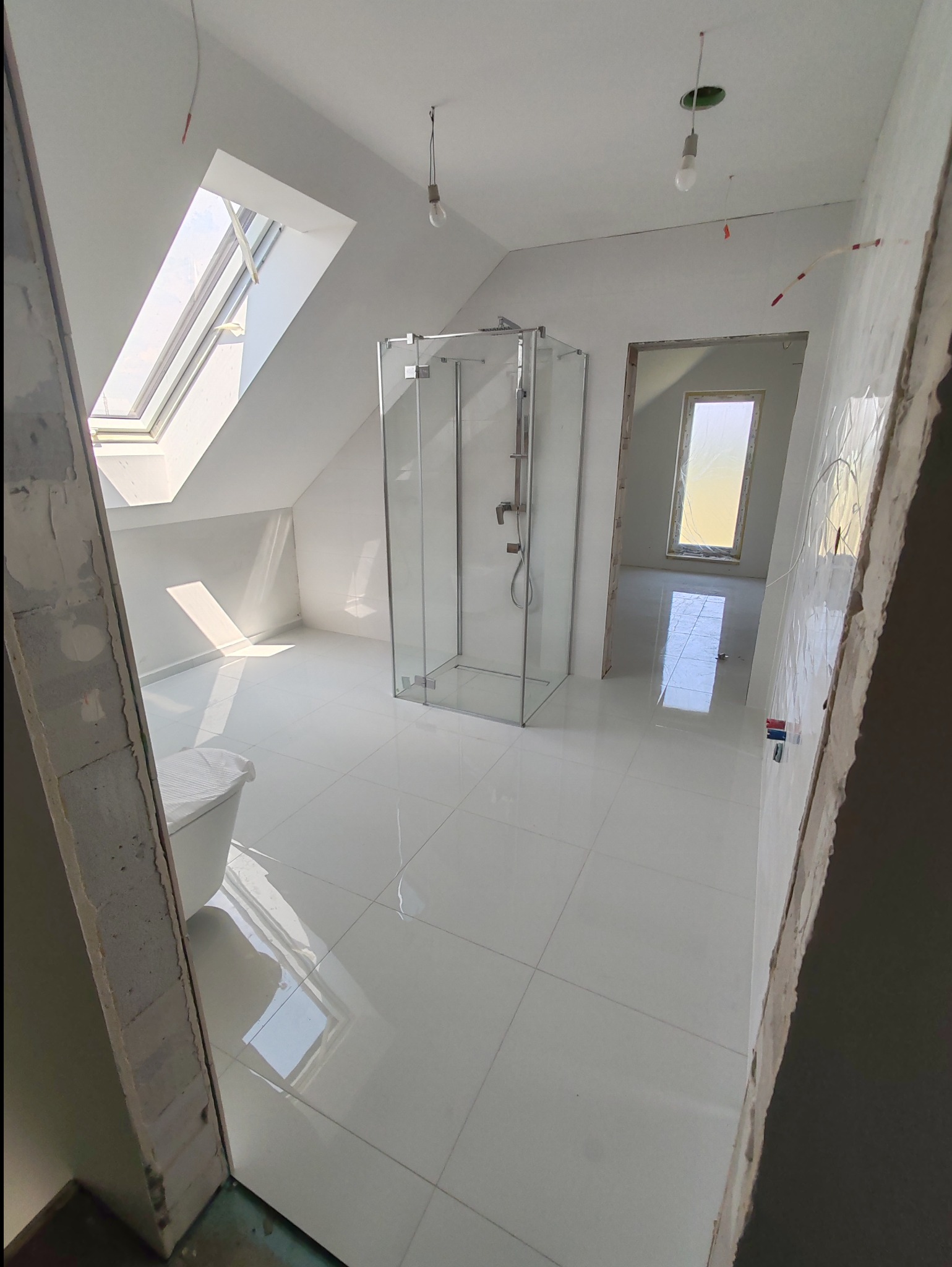 Łazienka na poddaszu w trakcie wykańczania: białe płytki podłogowe, kabina prysznicowa ze szkła, okno dachowe wpuszczające światło, surowy stan ścian z widocznymi instalacjami, widok na kolejne...