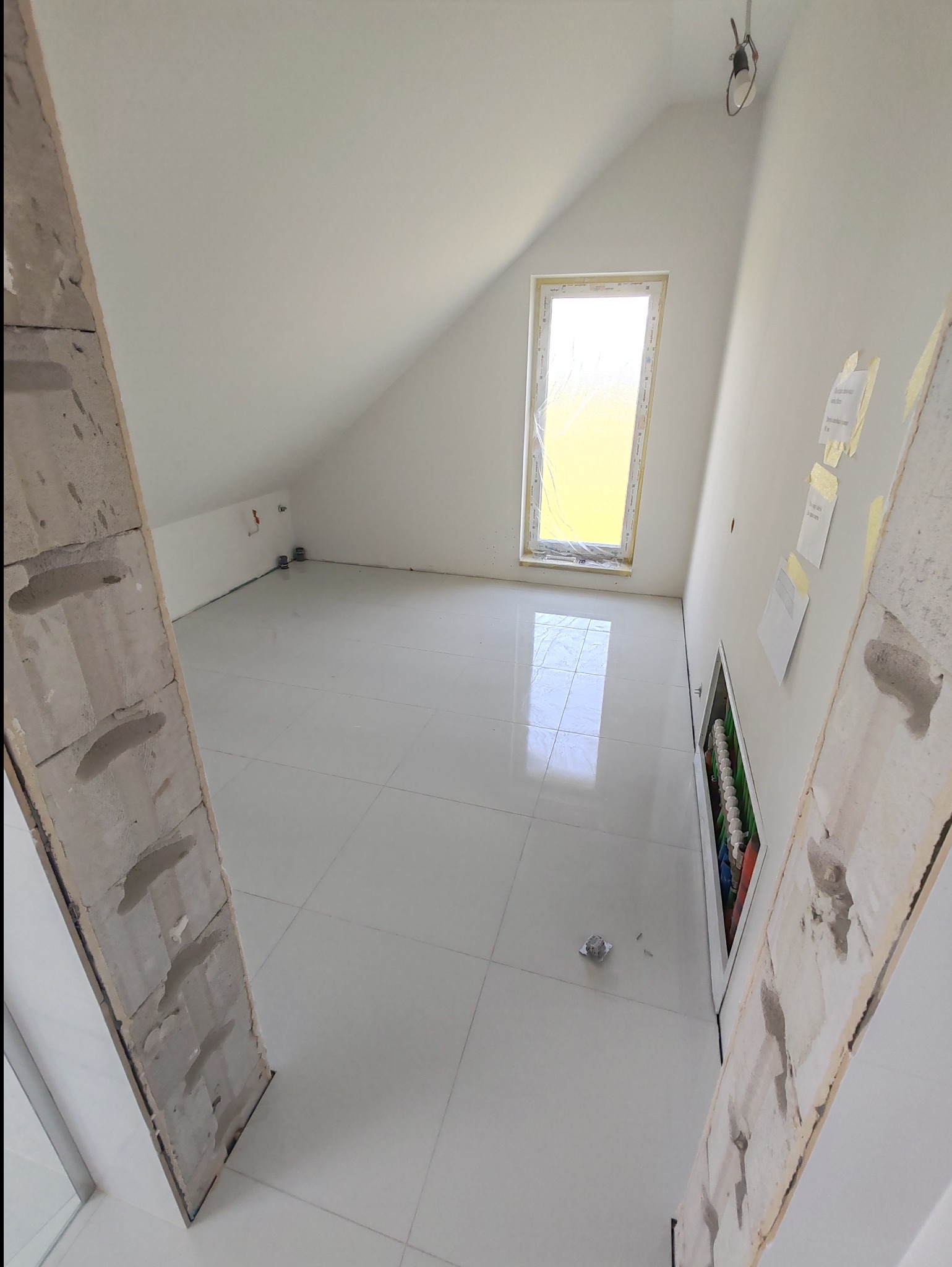 Poddasze z białymi płytkami podłogowymi, oknem połaciowym zabezpieczonym folią i fragmentem instalacji elektrycznej w ścianie, widok przez surową ościeżnicę.