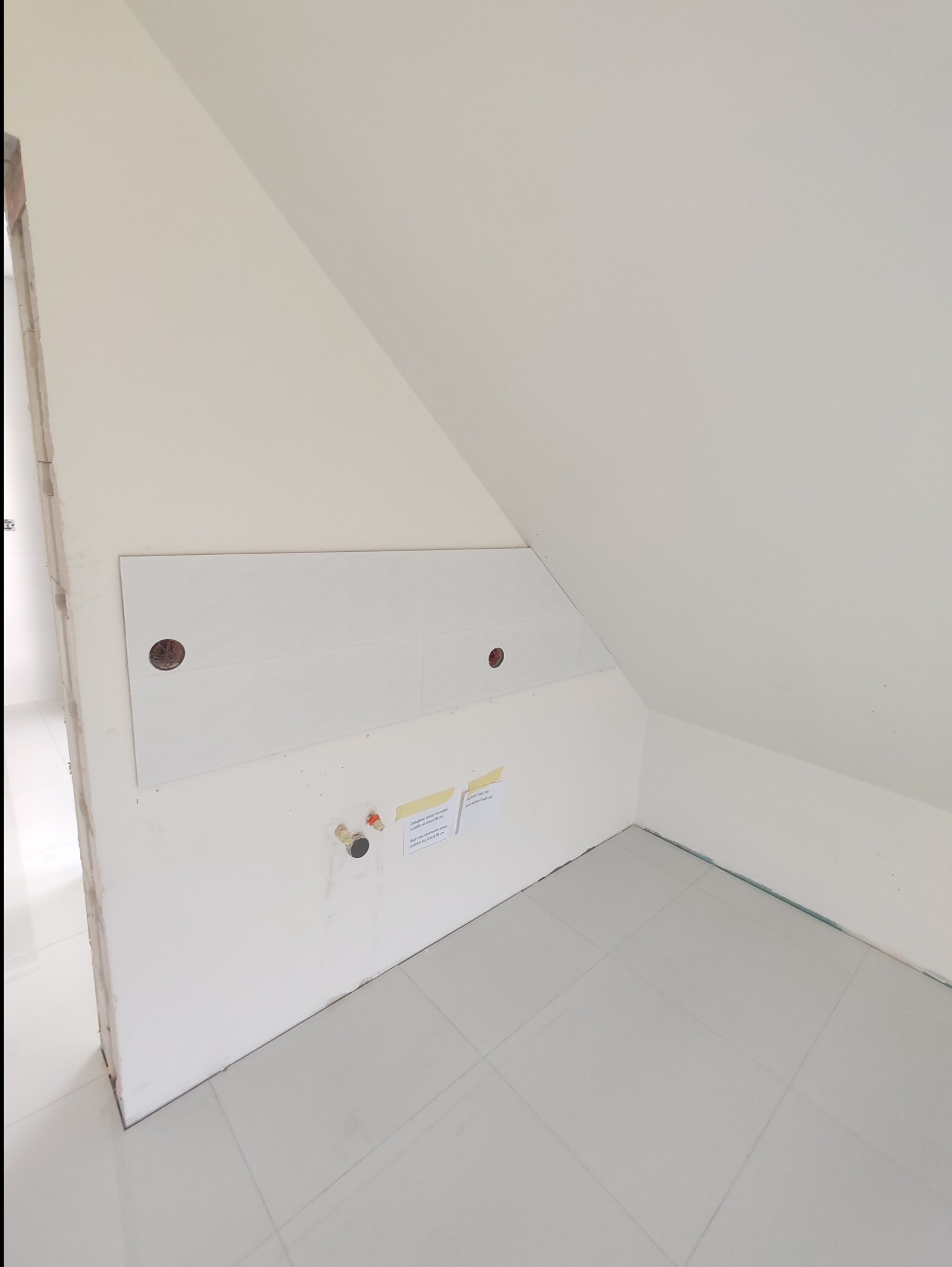 Białe płytki częściowo ułożone na ścianie w skosie poddasza, widoczne otwory na instalacje i notatki na ścianie.