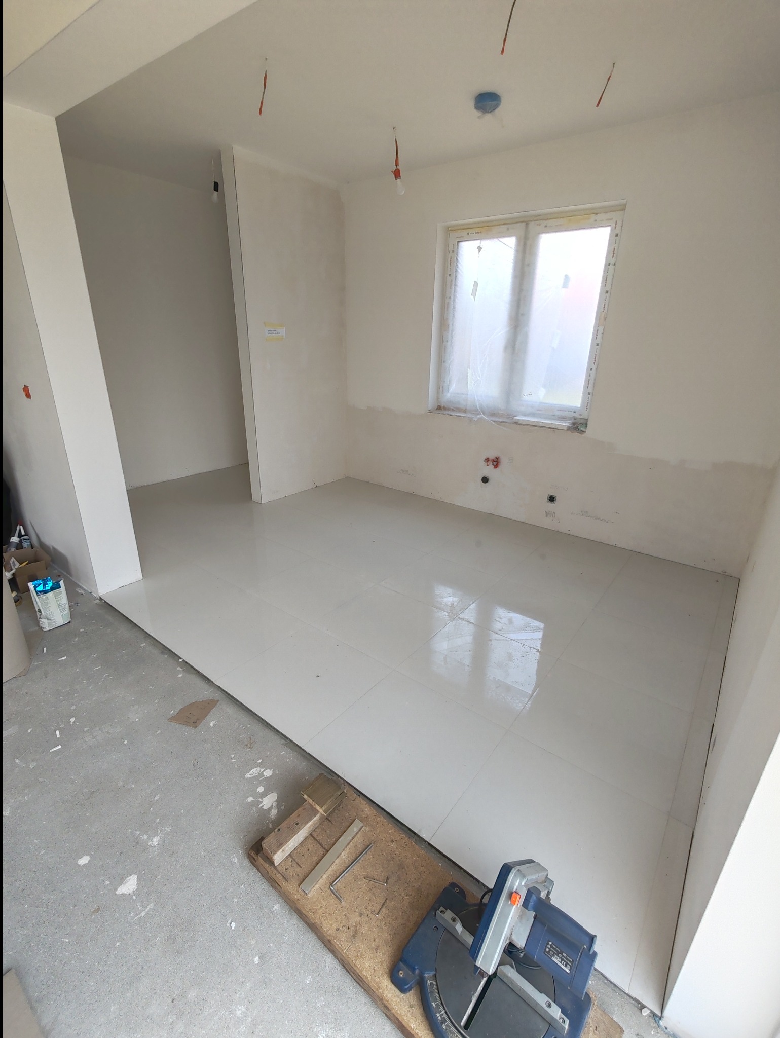 Białe, błyszczące płytki podłogowe ułożone w jasnym pomieszczeniu, widoczna piła do cięcia płytek na desce, okno zasłonięte folią malarską, przygotowane do fugowania.