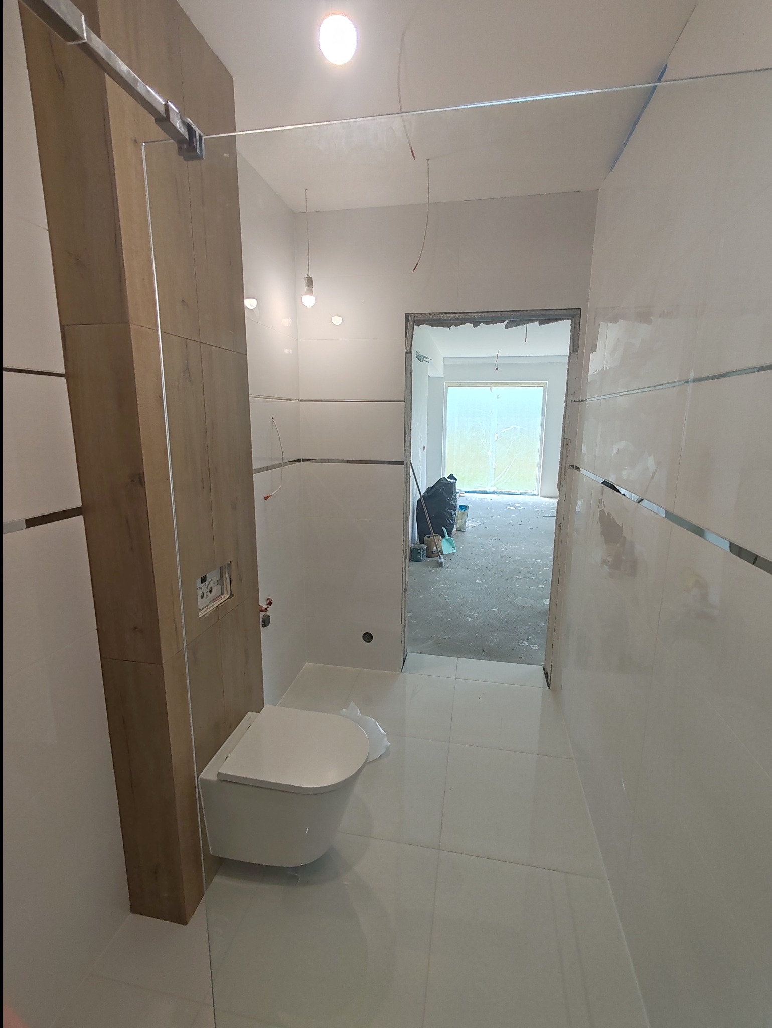 Nowoczesna łazienka w trakcie wykończenia: białe płytki, podwieszana toaleta, drewnopodobna zabudowa stelaża, widoczne niedokończone prace w sąsiednim pomieszczeniu.