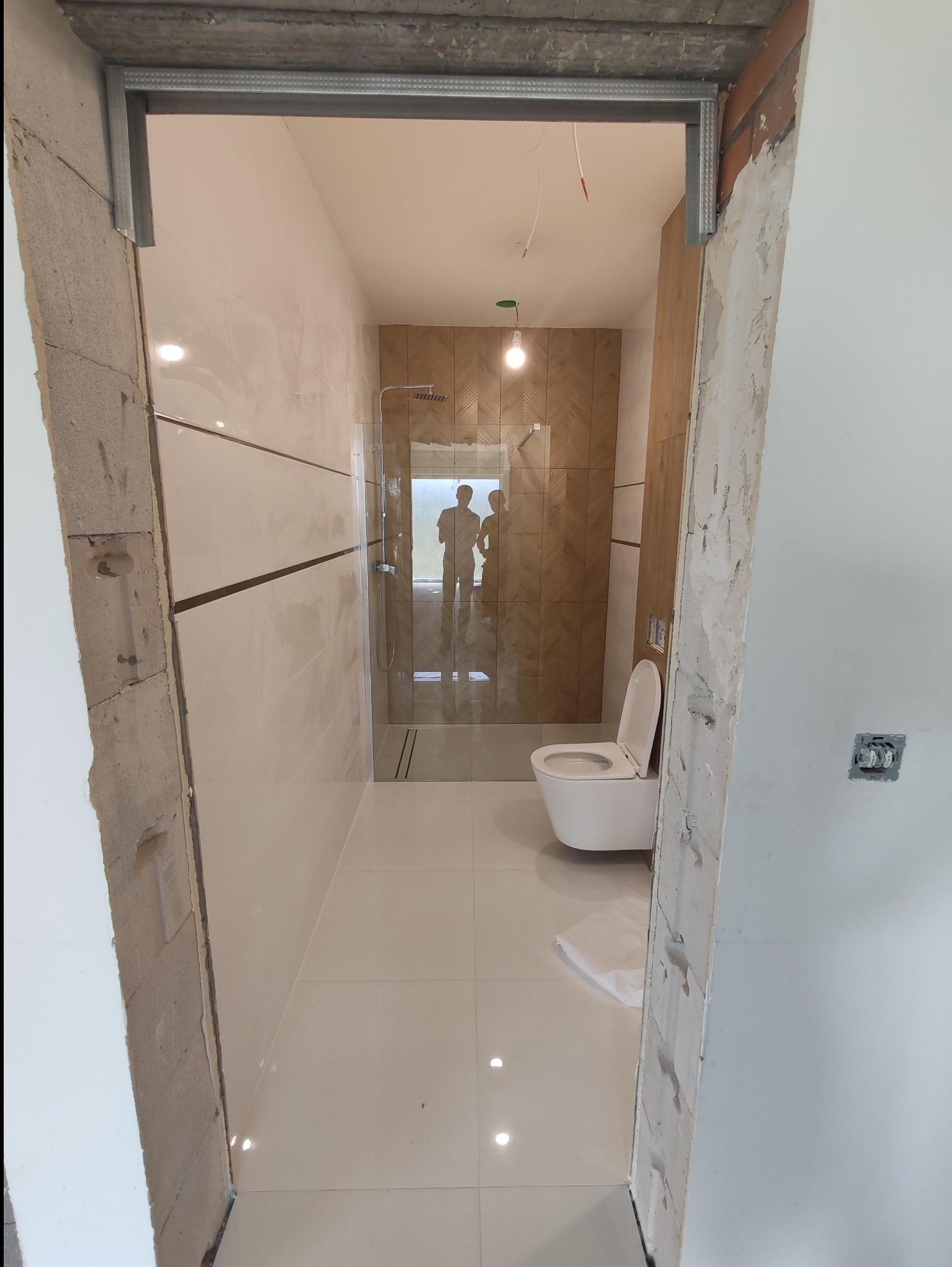 Wnętrze łazienki w trakcie remontu, widoczne białe płytki, toaleta wisząca i kabina prysznicowa z drewnopodobną ścianą w jodełkę, surowy otwór drzwiowy z metalową ramą.