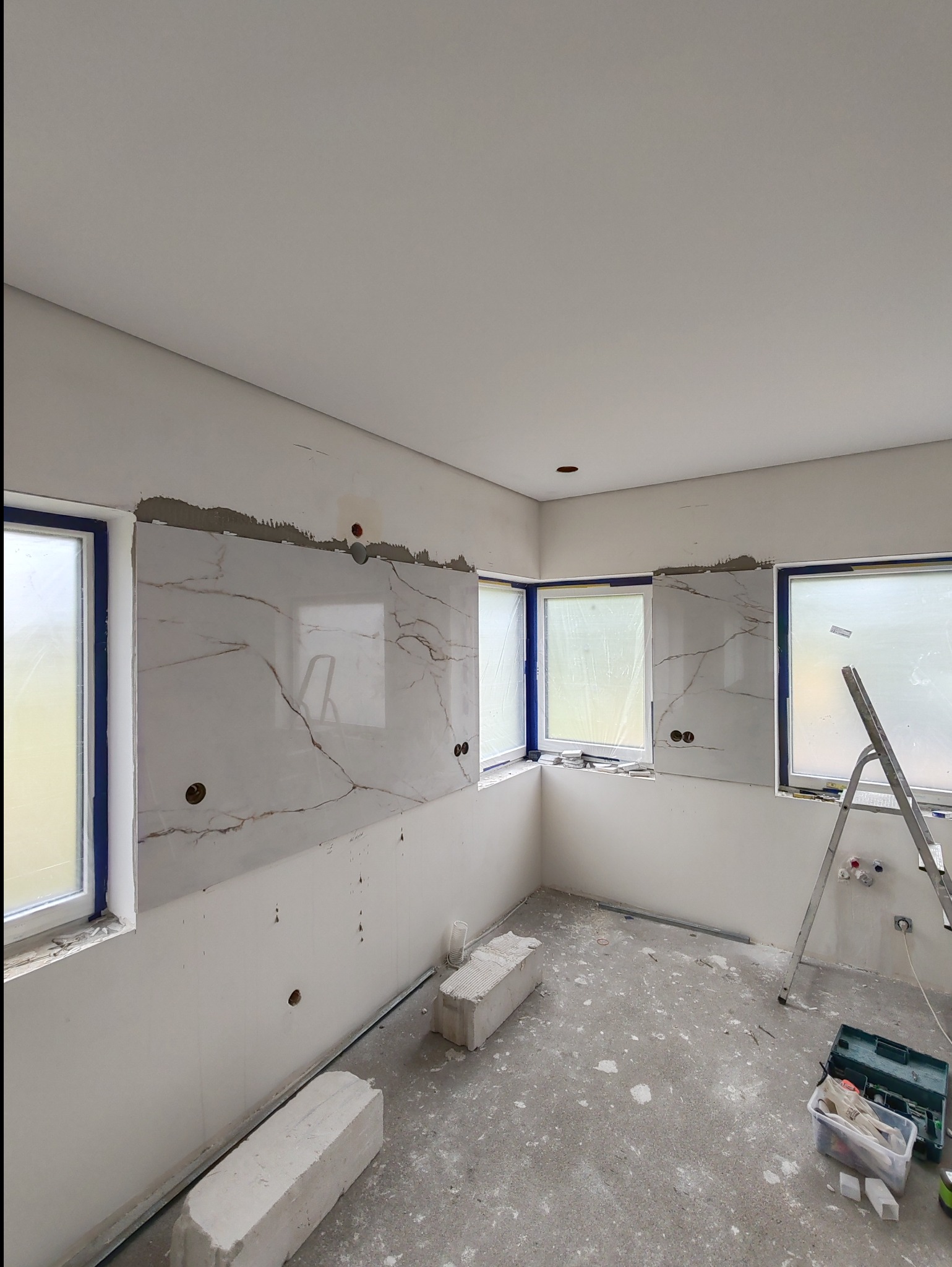 Montaż marmurowych płyt na ścianie w pomieszczeniu z trzema oknami, widoczne oklejone ramy okienne, drabina, resztki zaprawy i narzędzia na podłodze.