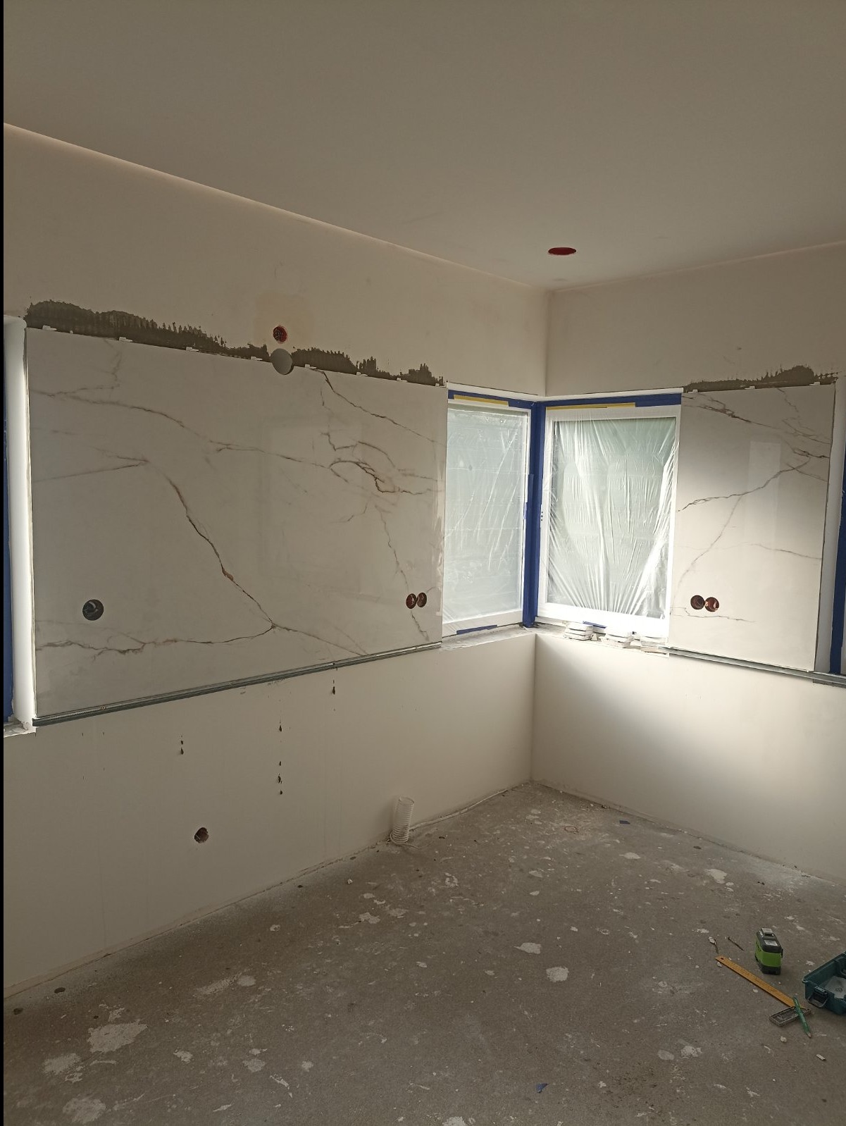 Wnętrze w trakcie montażu płytek imitujących marmur na ścianach, okna zabezpieczone folią, widoczne narzędzia pomiarowe na podłodze.