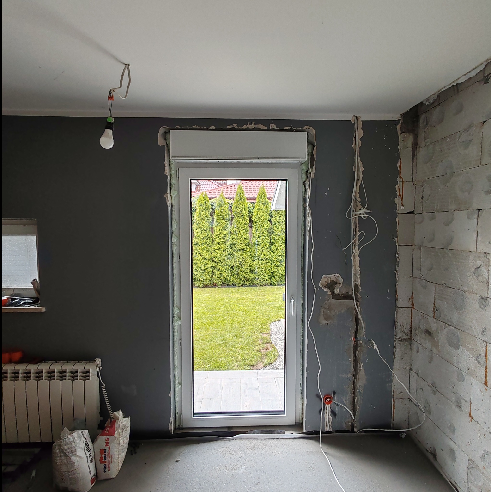 Wnętrze w trakcie remontu z nowo zamontowanym oknem z roletą zewnętrzną, widoczne przewody elektryczne, surowa ściana z betonu komórkowego, grzejnik i worki z zaprawą.