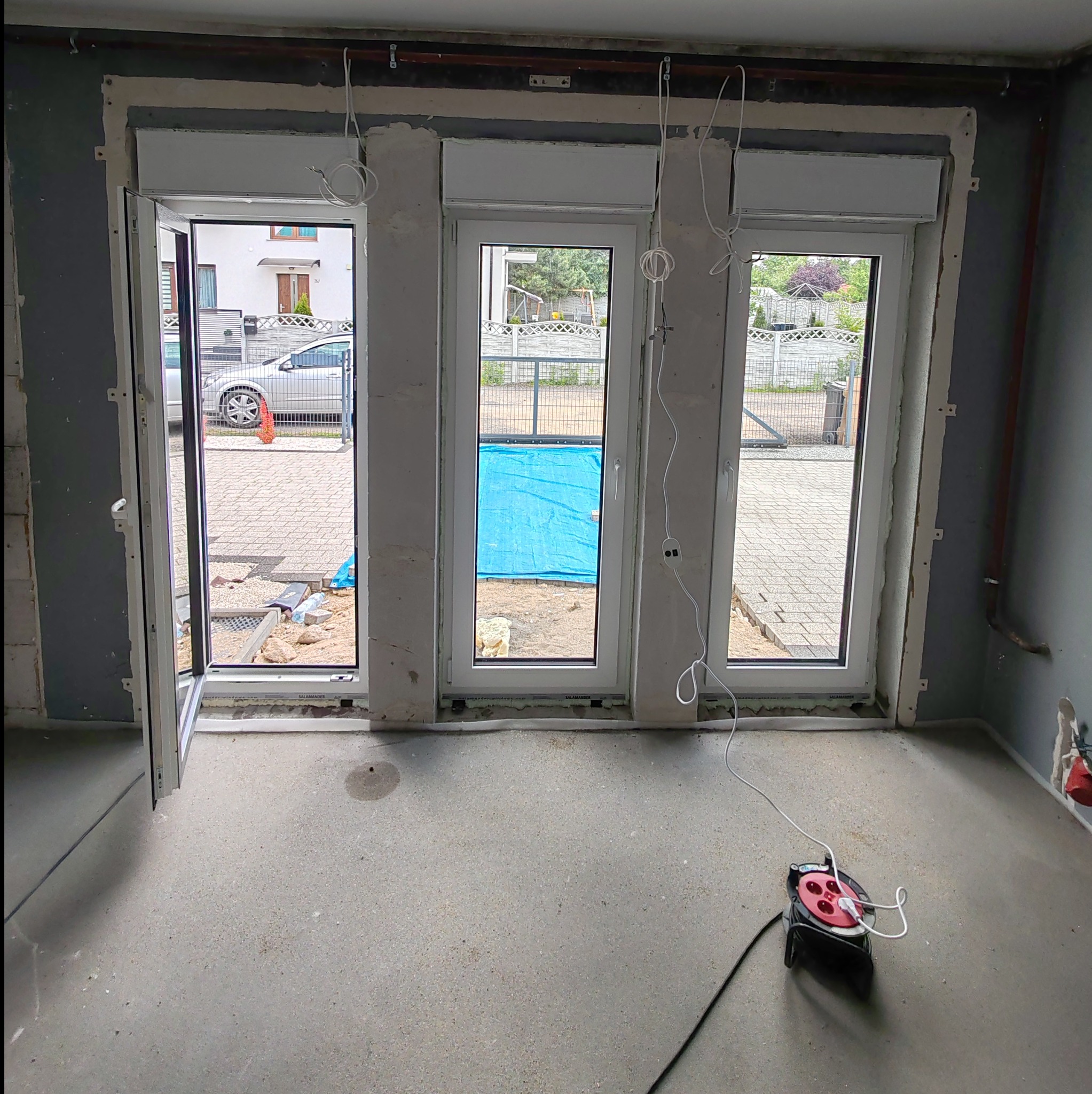 Montaż okien: trzy białe okna z roletami zewnętrznymi w surowym stanie, jedno okno otwarte, widoczne przewody elektryczne i przedłużacz na podłodze.