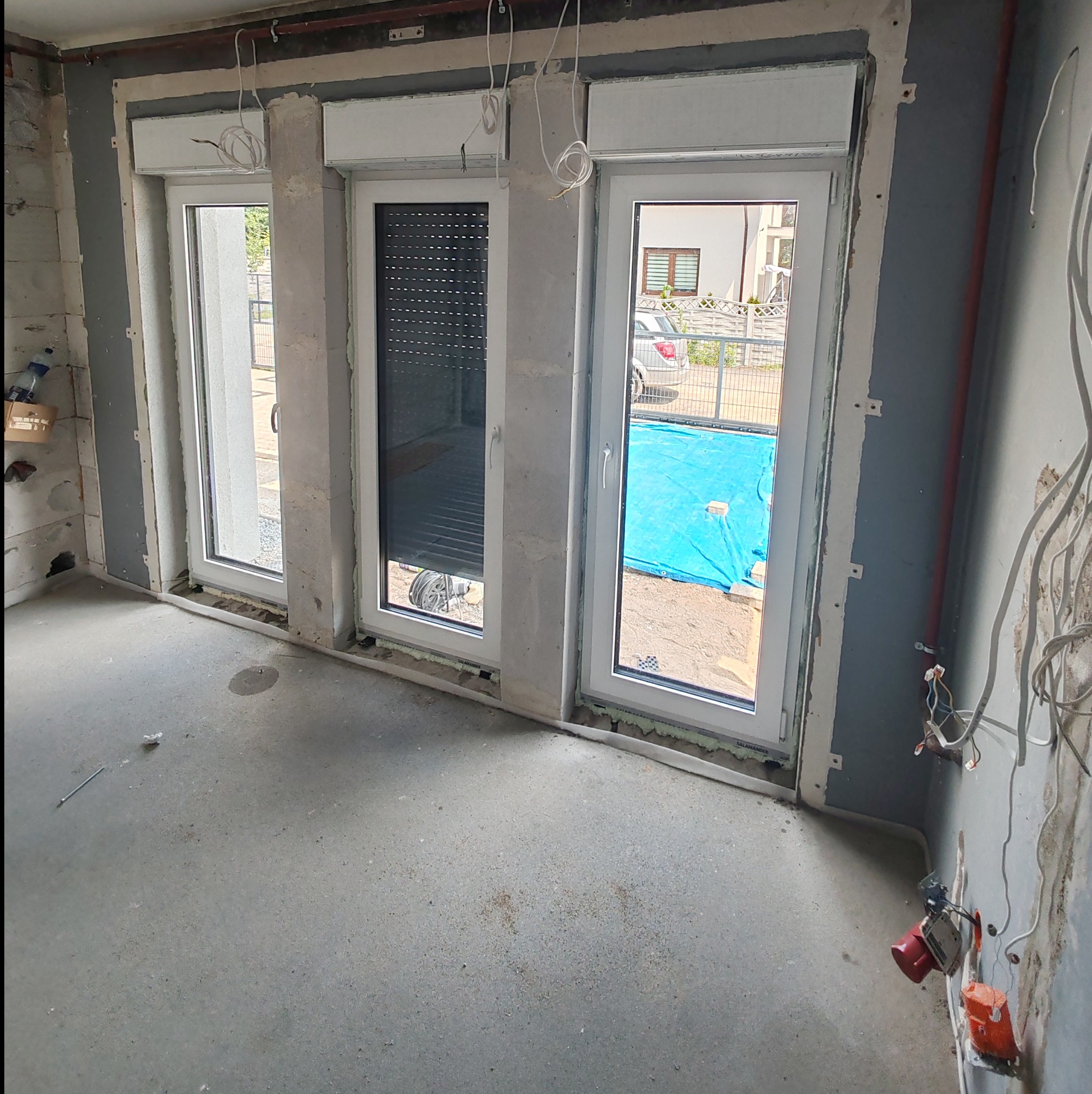 Wnętrze w trakcie remontu z trzema oknami z roletami zewnętrznymi, widoczne ślady prac wykończeniowych na ścianach i podłodze, instalacja elektryczna w trakcie montażu.