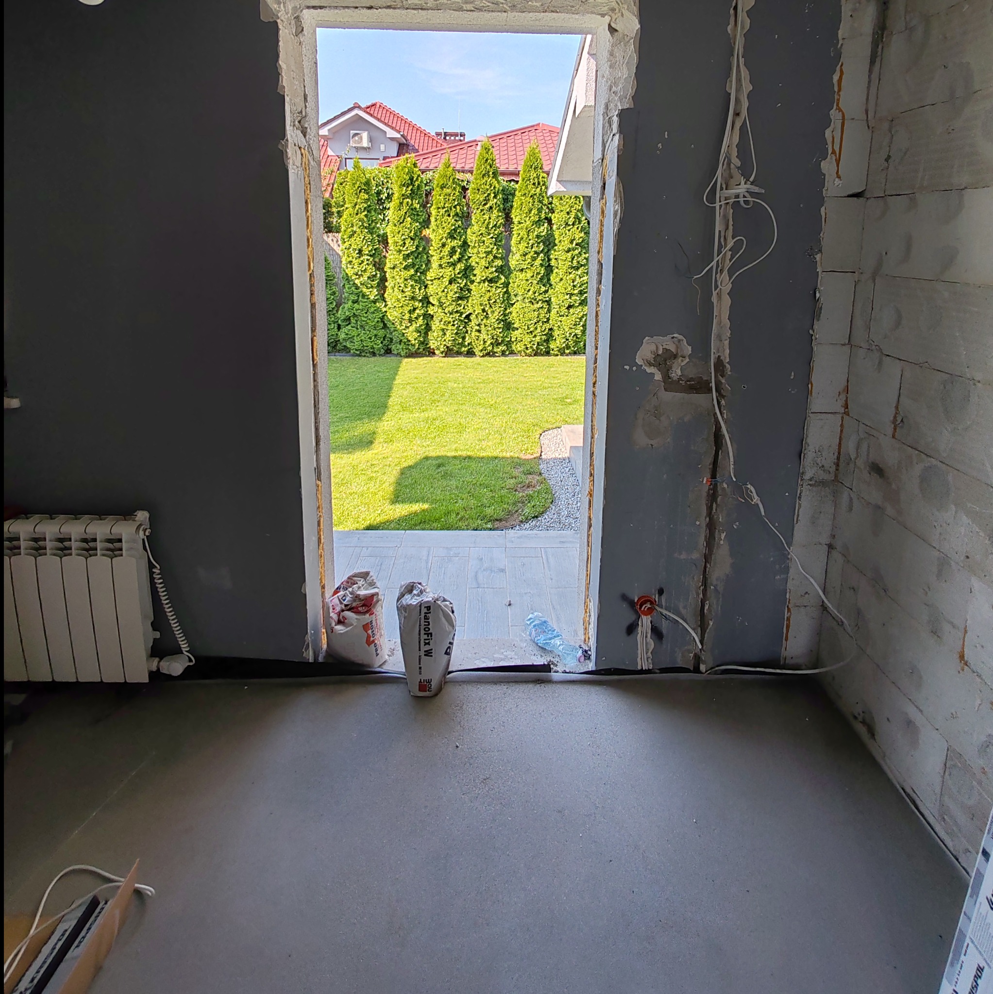 Przygotowane podłoże pod wylewkę w pomieszczeniu w trakcie remontu, widoczne worki z zaprawą i instalacja elektryczna, z widokiem na ogród przez otwór drzwiowy.