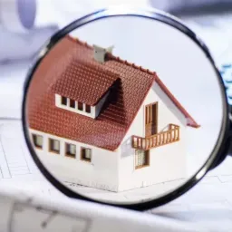 Model domu z czerwoną dachówką oglądany przez szkło powiększające, leżący na planach architektonicznych obok kalkulatora i zwiniętych rysunków.