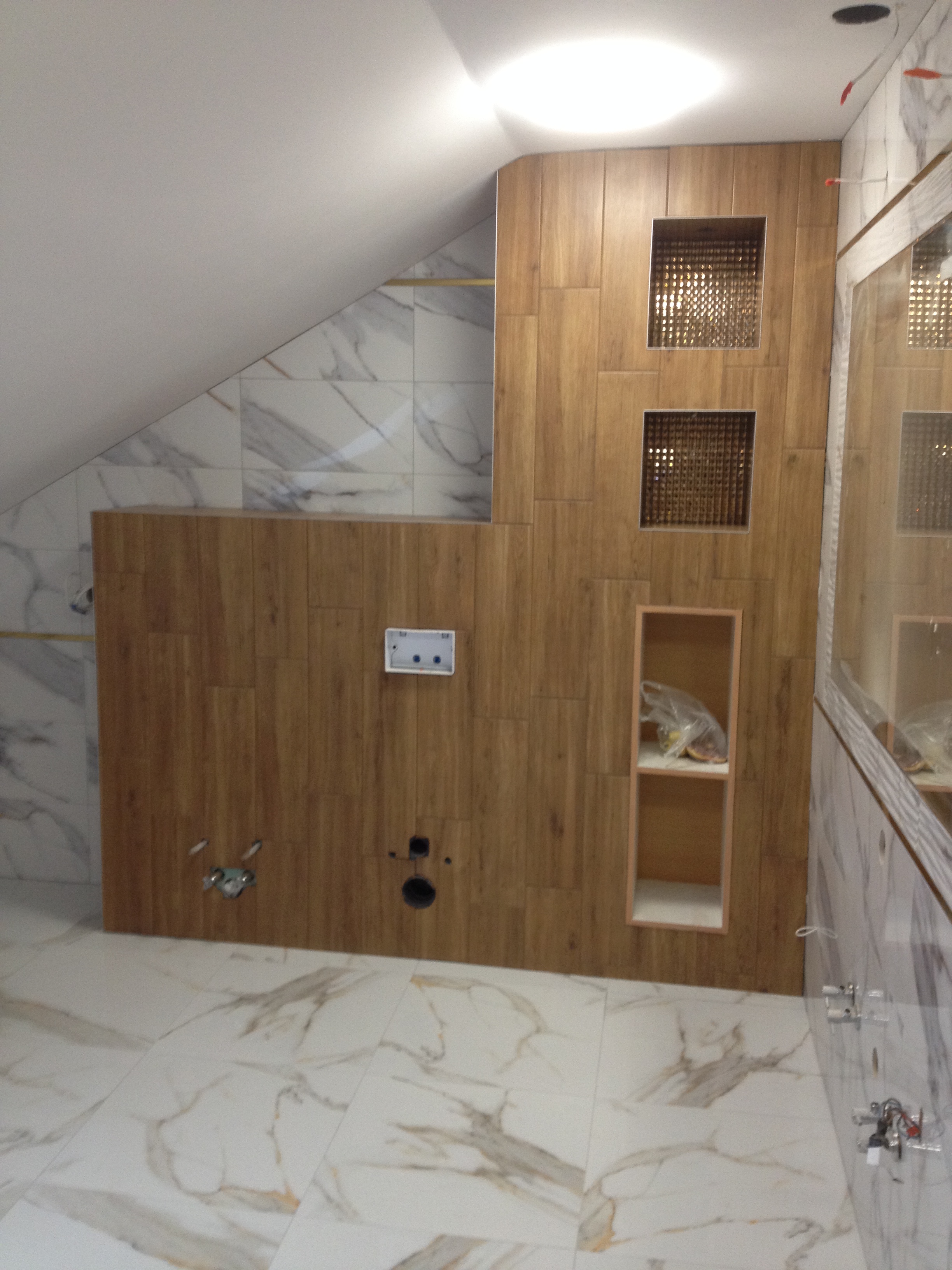 Wnętrze łazienki w trakcie prac wykończeniowych, widoczne drewnopodobne płytki na ścianach, marmurowe płytki podłogowe, przygotowane otwory na armaturę i wnęki dekoracyjne, oraz instalacja...