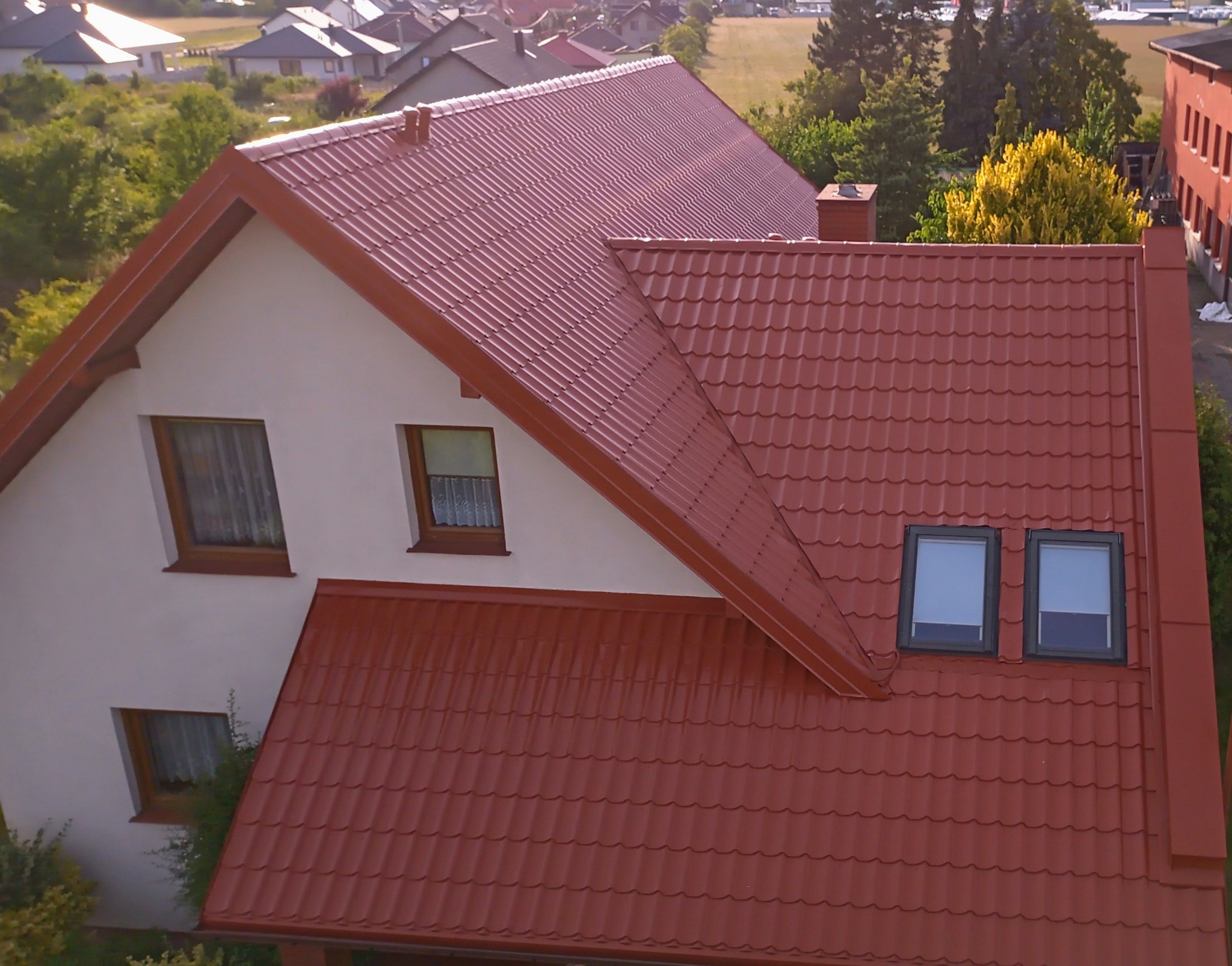 Widok z góry na dom z nowym, czerwonym dachem z blachodachówki, z dwoma oknami dachowymi, na tle zabudowy jednorodzinnej i zieleni.