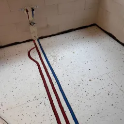 Czerwona i niebieska rura instalacji grzewczej ułożona na białej izolacji podłogowej w surowym wnętrzu, widoczne mocowania na ścianie z betonu komórkowego.