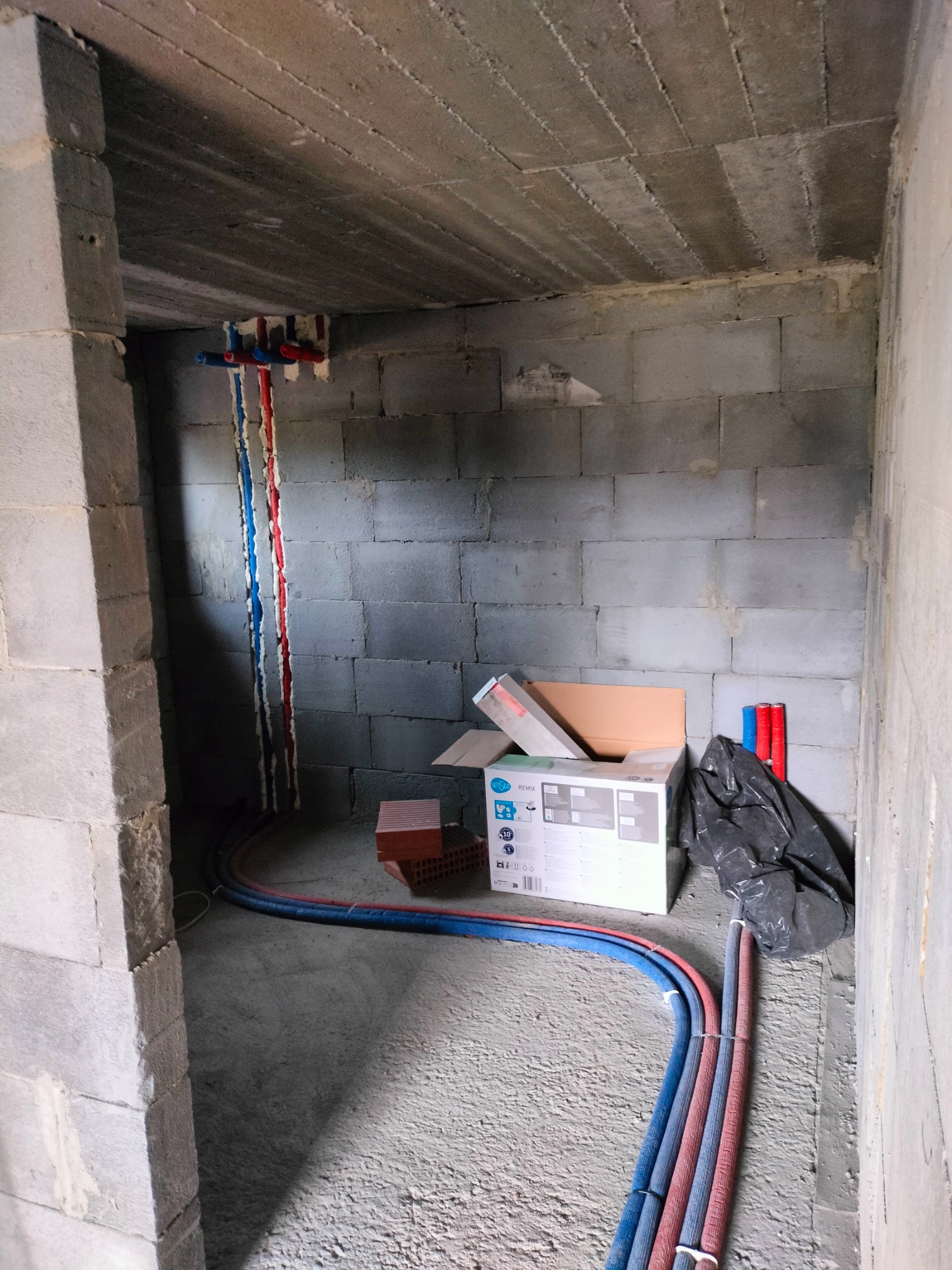 Surowy stan instalacji w pomieszczeniu z betonowych bloczków: rury ciepłej i zimnej wody w izolacji prowadzone wzdłuż ściany, wystające przyłącza, kartonowe opakowanie armatury i gruz na podłodze.