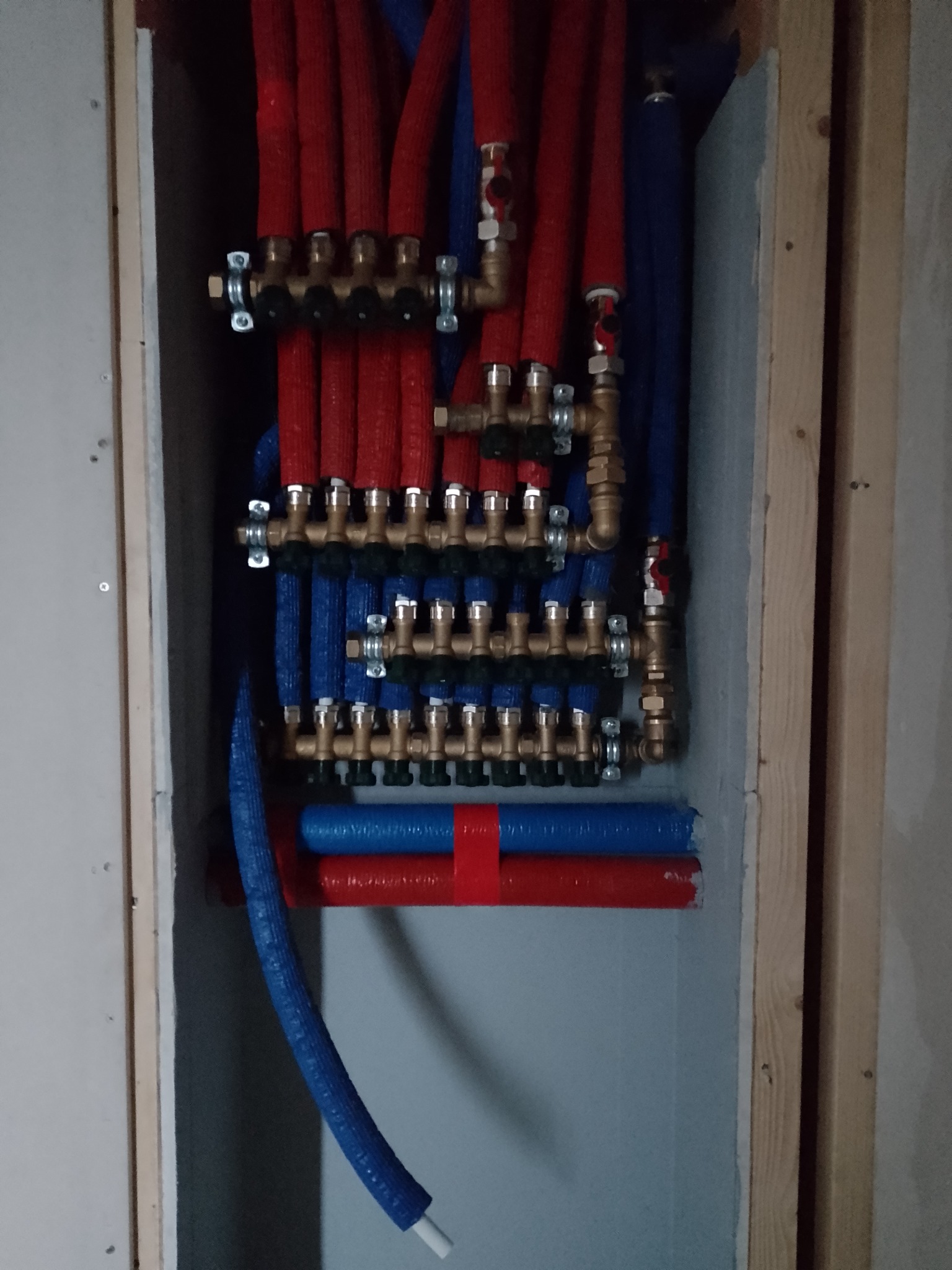 Rozdzielacz instalacji grzewczej i wodnej z rurami w izolacji, zamontowany w surowej wnęce z drewnianą ramą, widoczne liczne zawory i złączki.