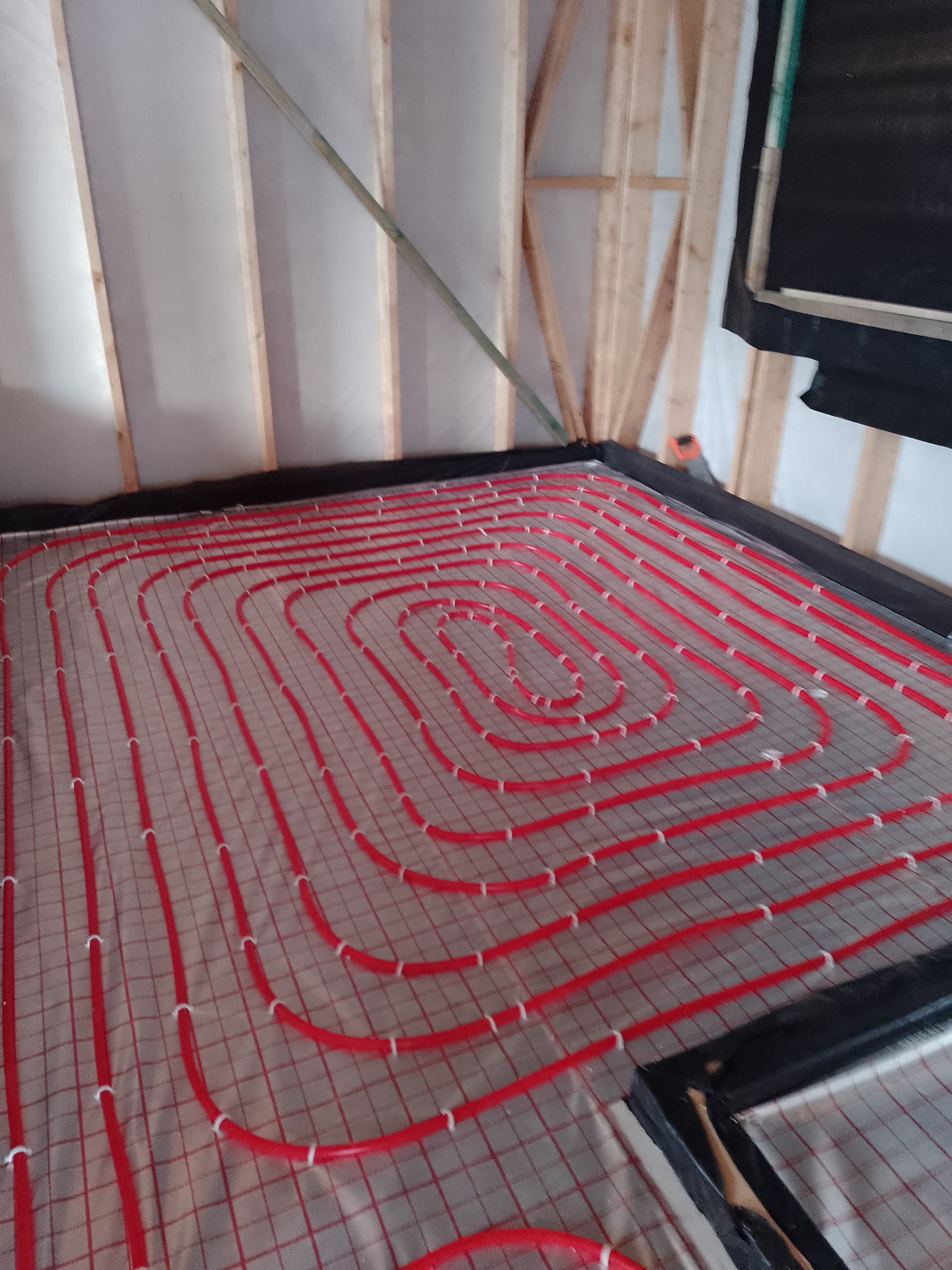 Czerwone rurki ogrzewania podłogowego ułożone spiralnie na siatce, widoczne elementy konstrukcji drewnianej ściany i izolacja.