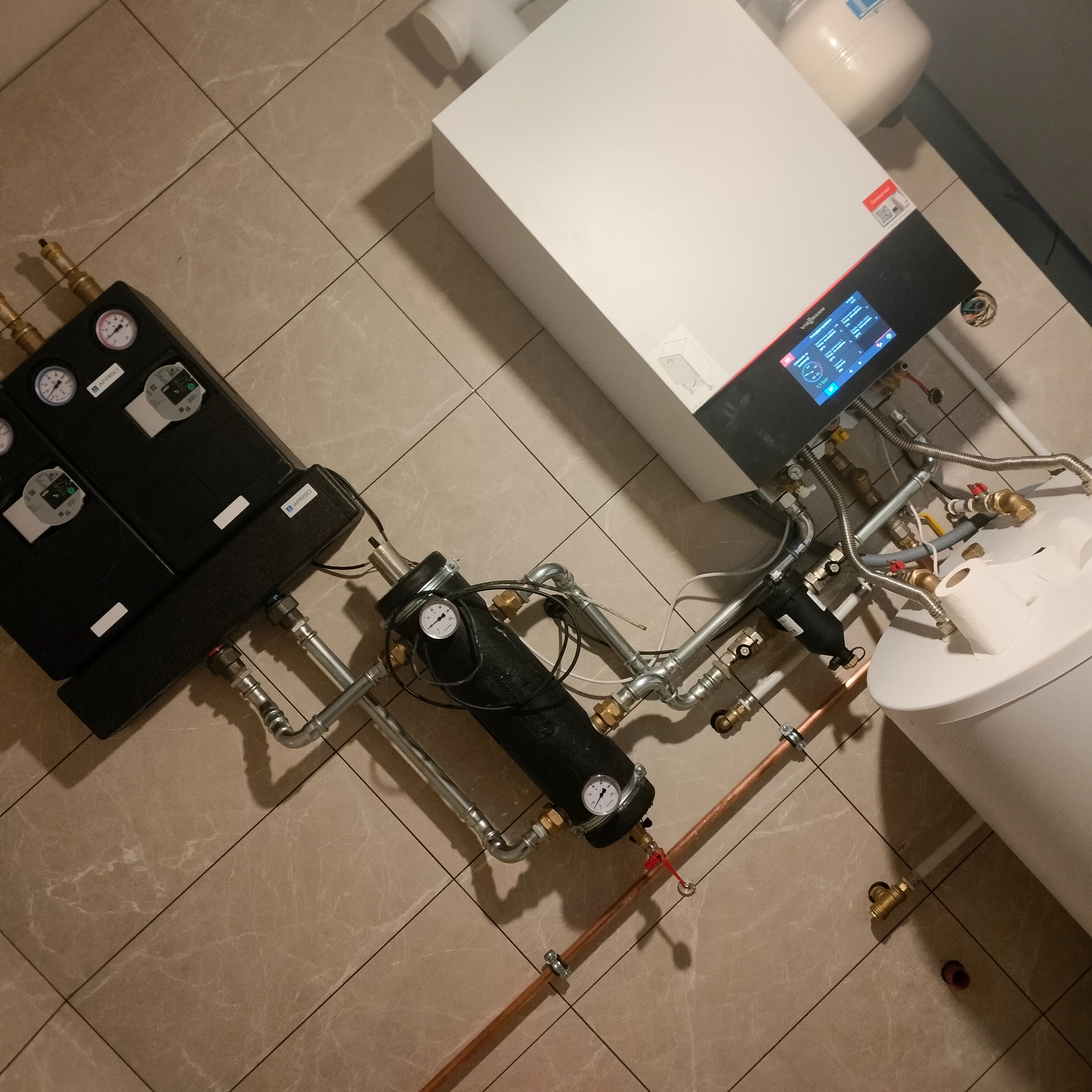 Nowoczesny piec centralnego ogrzewania z czarnymi modułami sterującymi i rurami miedzianymi, zainstalowany na płytkach podłogowych, widoczne manometry i elementy przyłączeniowe.