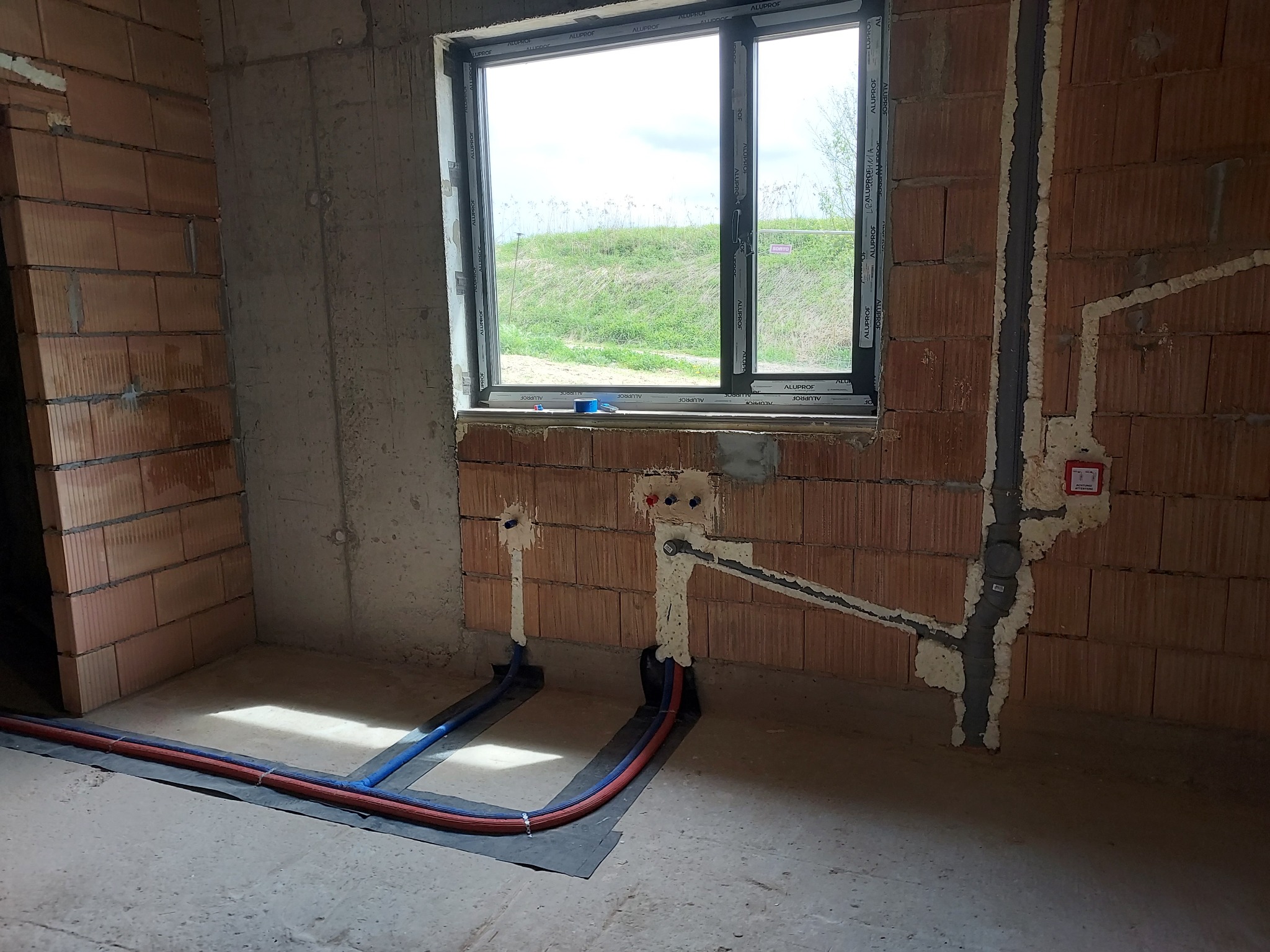 Surowy stan instalacji wodnej i kanalizacyjnej w budynku z cegły, z widocznymi rurami w ścianach i podłodze, zabezpieczonymi pianką montażową, oraz oknem z widokiem na zieleń.