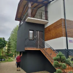 Malowanie fragmentu elewacji budynku z balkonem i schodami zewnętrznymi; widoczny pracownik w trakcie pracy, malujący czarną farbą dolną część elewacji.