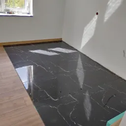 Ułożona, błyszcząca, czarna podłoga z marmurowym wzorem w nowym pomieszczeniu z białymi ścianami, widoczne niedokończone listwy przypodłogowe i resztki materiałów budowlanych.