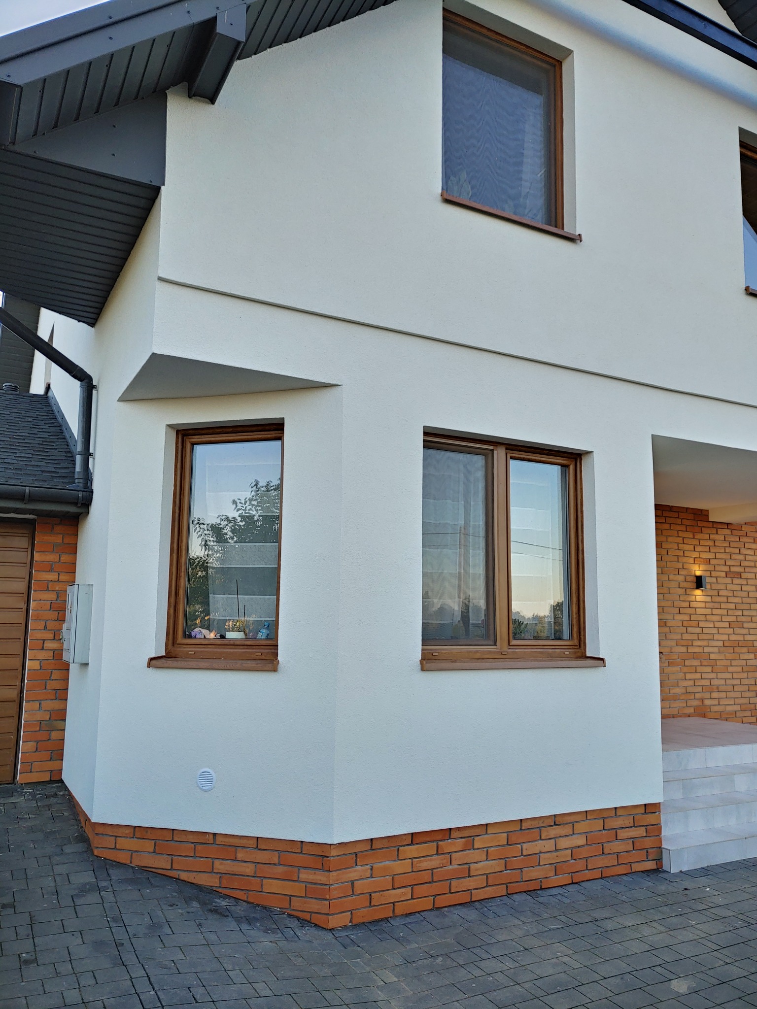 Dwukondygnacyjny dom z białą elewacją, brązowymi ramami okiennymi i ceglaną podmurówką, widoczny z zewnątrz na tle kostki brukowej.