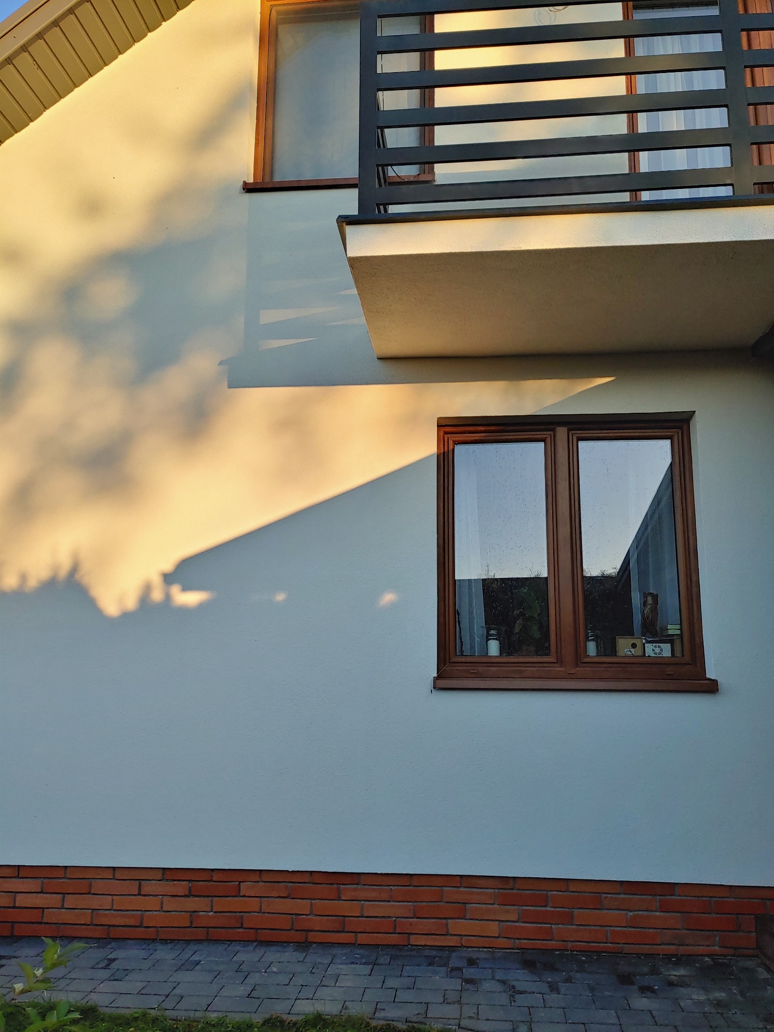 Fragment elewacji domu z oknem z brązową ramą i balkonem, oświetlony popołudniowym słońcem, rzucającym cienie drzew na białą ścianę nad czerwoną ceglaną podmurówką.