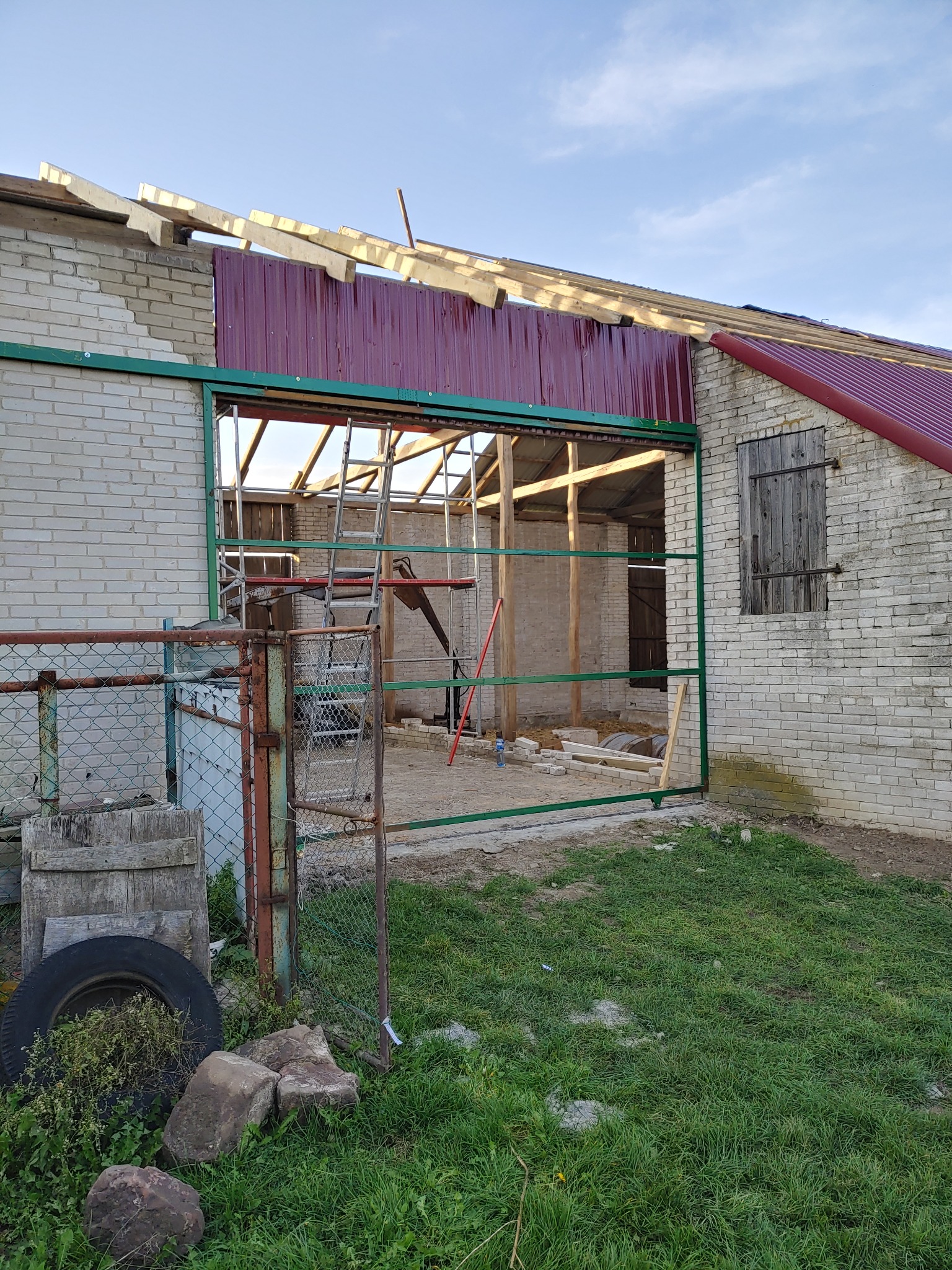 Konstrukcja budynku gospodarczego w Waśniowie z częściowo zamontowanym dachem z blachy trapezowej w kolorze bordowym, widoczna drabina i drewniane elementy więźby dachowej.
