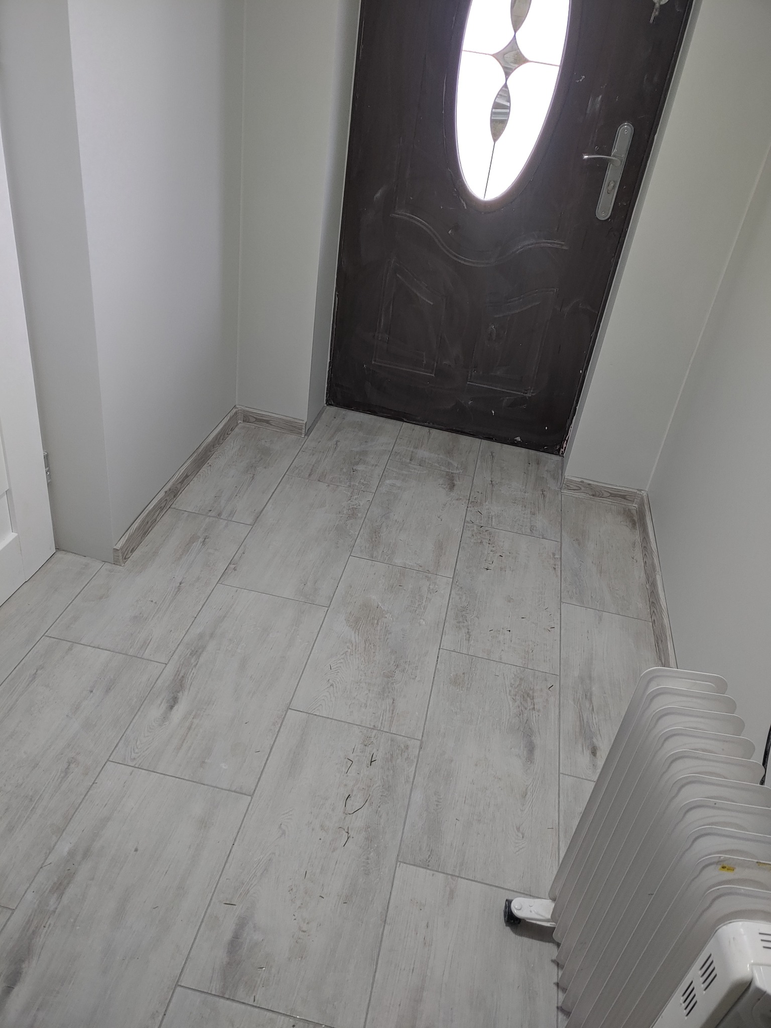 Wnętrze korytarza z szarą podłogą imitującą drewno, ciemnymi drzwiami z owalnym przeszkleniem i białymi ścianami, widoczne ślady po remoncie na podłodze, w rogu elektryczny grzejnik.