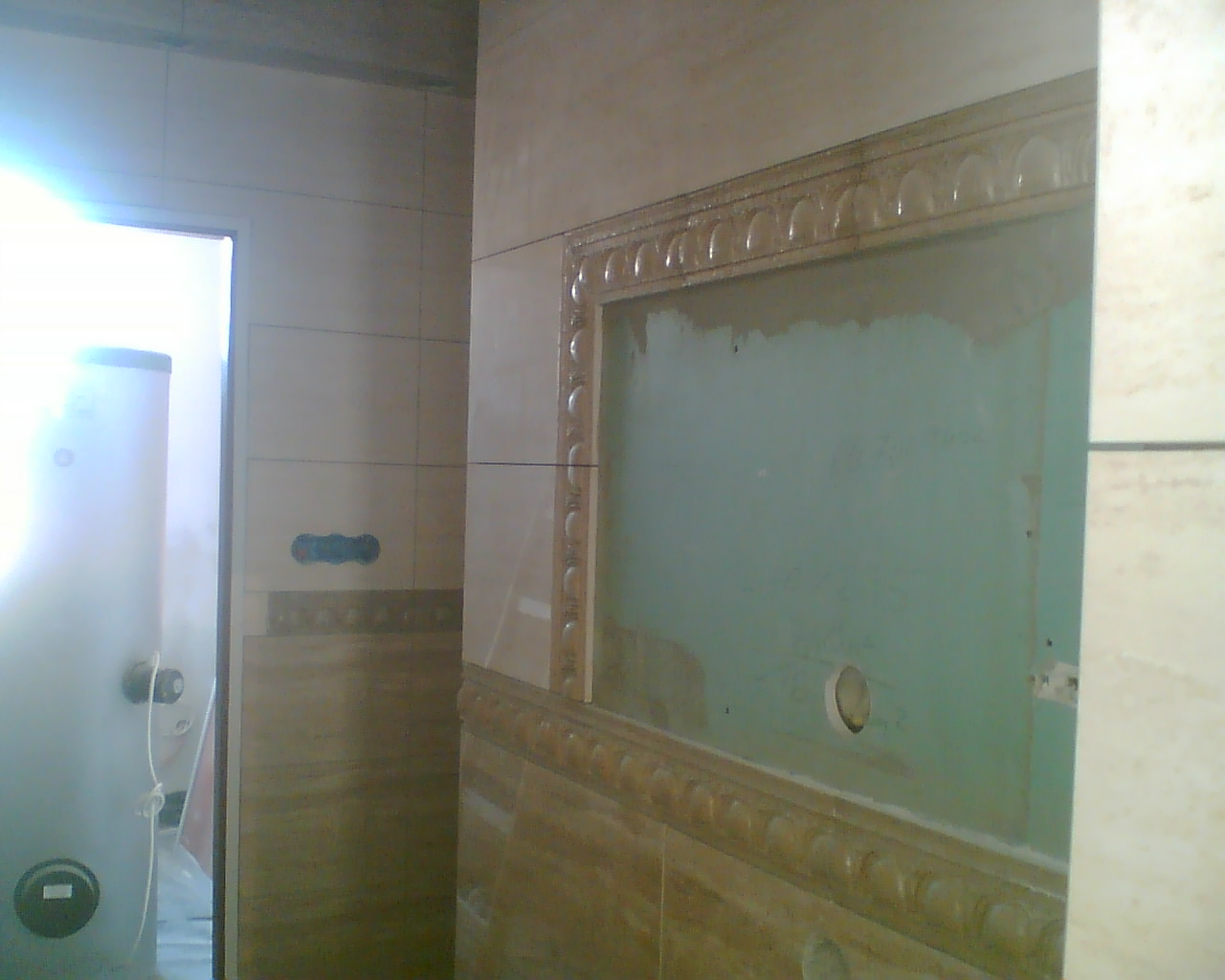 Wnętrze w trakcie remontu: ułożone płytki imitujące drewno z dekoracyjnym obramowaniem wokół wnęki, widoczny podgrzewacz wody w tle.