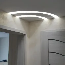 Ściana z dekoracyjną półką pod sufitem, podświetloną taśmą LED, widoczne dwie pary drzwi w kolorze jasnego drewna z poziomymi, ciemnymi intarsjami.