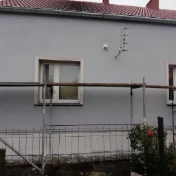 Świeżo pomalowana szara elewacja domu jednorodzinnego z białymi obramowaniami okien, widoczne rusztowanie, antena na ścianie i fragment ogrodu z kwitnącą różą.