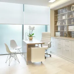 Jasny gabinet lekarski z biurkiem, krzesłami, kwiatami i zabudową meblową z przeszkloną witryną.