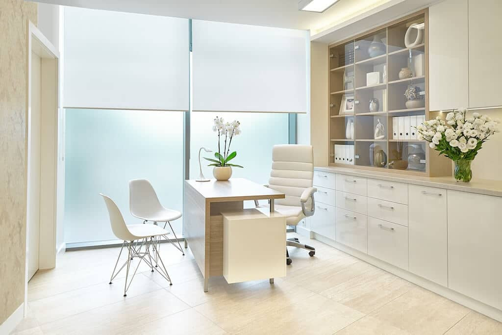 Jasny gabinet lekarski z biurkiem, krzesłami, kwiatami i zabudową meblową z przeszkloną witryną.
