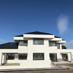 Nowoczesny dom z płaskim dachem i dużymi oknami, jeszcze z folią ochronną, w trakcie wykończenia terenu wokół budynku.