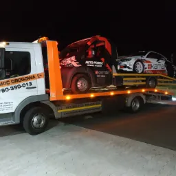 Laweta z białym i pomarańczowym nadwoziem przewozi uszkodzony czarny bus i białe auto sportowe z naklejkami, nocne ujęcie.