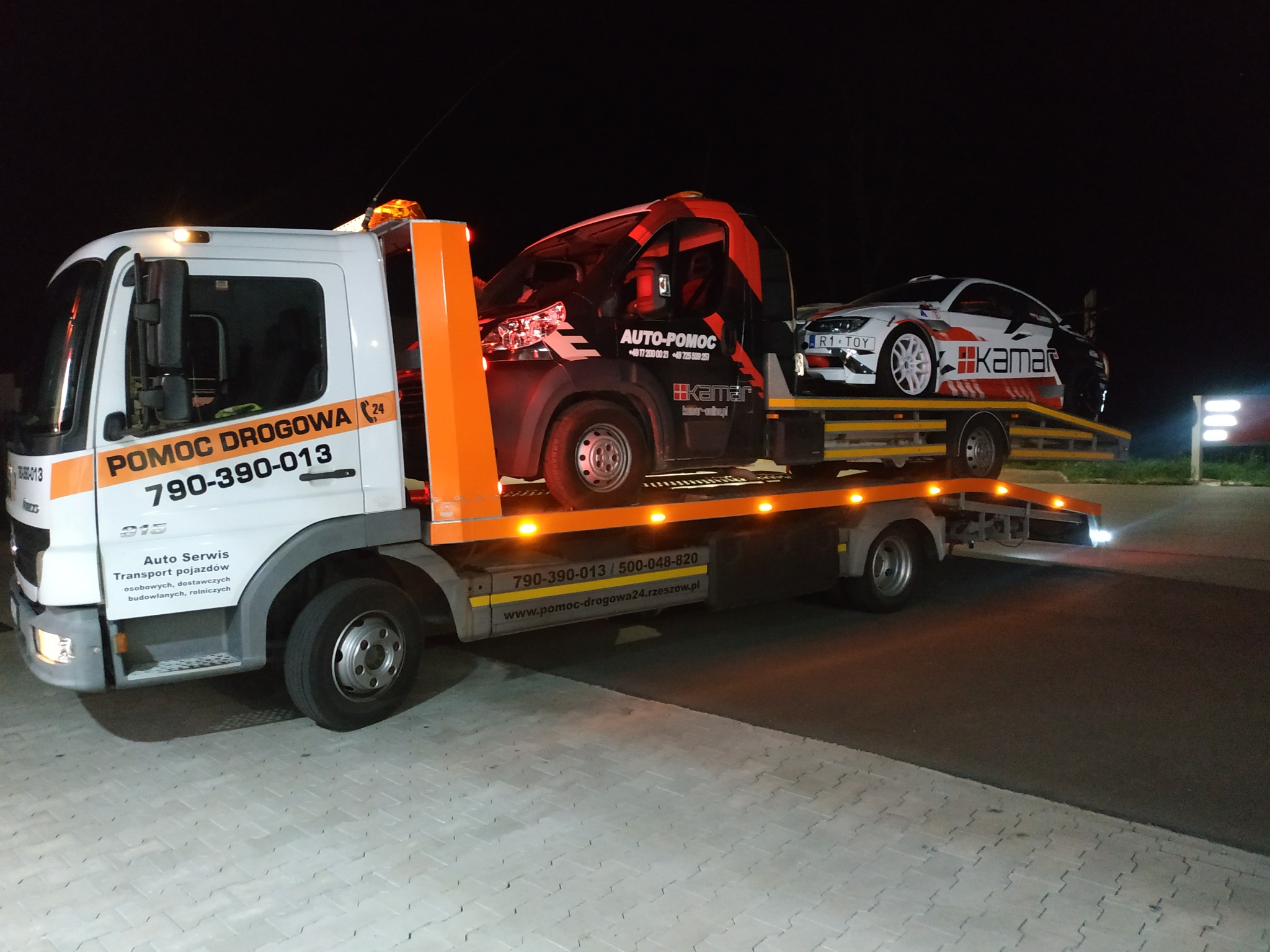 Laweta z białym i pomarańczowym nadwoziem przewozi uszkodzony czarny bus i białe auto sportowe z naklejkami, nocne ujęcie.