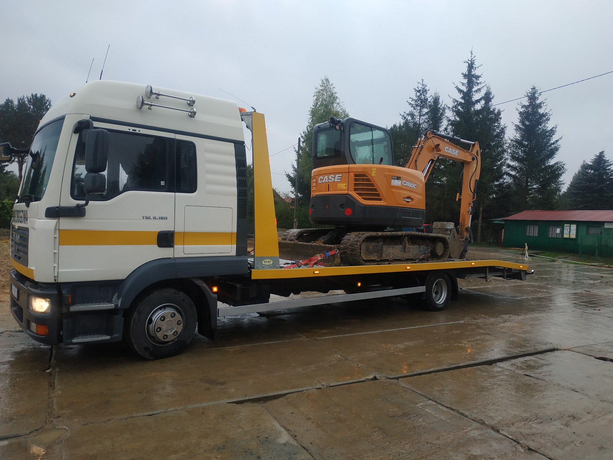 Biała laweta MAN TGL 8.180 z żółtym pasem transportuje pomarańczową koparkę CASE CX60C, zabezpieczoną czerwonymi pasami, na mokrym betonowym placu.