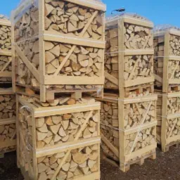 Stosy drewnianych skrzyń wypełnionych pociętym drewnem kominkowym na paletach, zabezpieczonych folią, na zewnątrz.