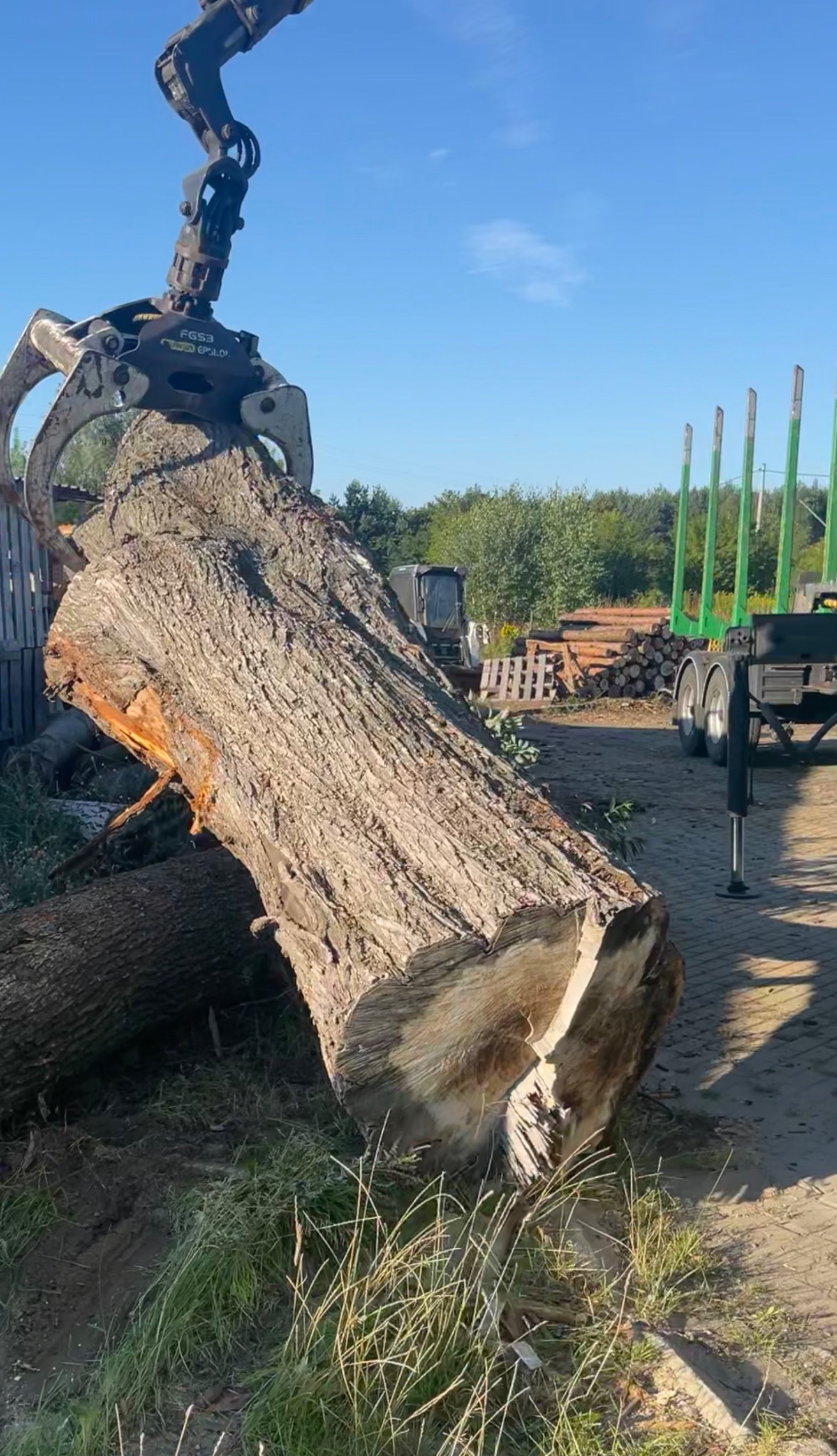 Manipulator chwytakowy podnosi kłodę drewna na placu składowym, widoczne stosy drewna w tle.