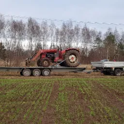 Czerwony traktor z ładowaczem na niskopodwoziowej przyczepie, transportowany białym busem na tle pola i drzew.